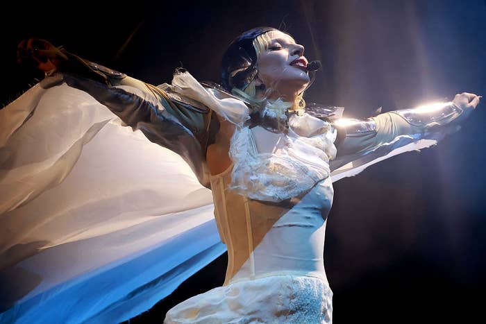 Performer on stage with arms outstretched, wearing an avant-garde costume featuring sheer fabric and structured details