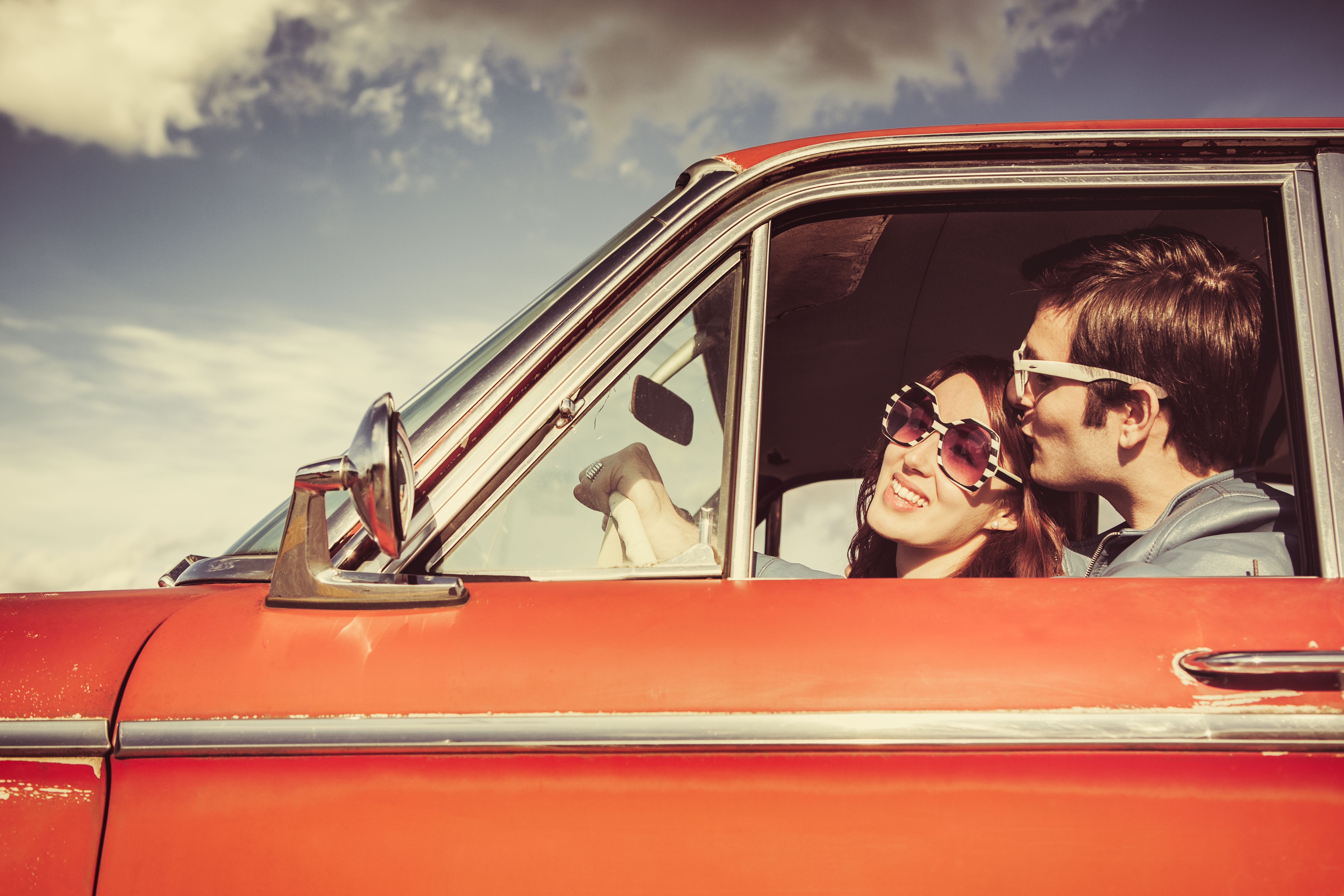 A couple wearing sunglasses smiles and enjoys a drive in a classic car, embodying a carefree and romantic vibe
