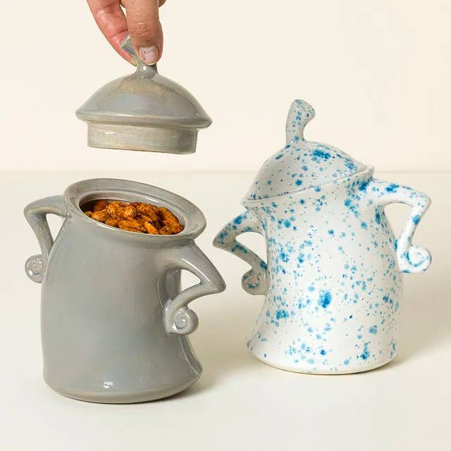 Two whimsical ceramic jars with curled handles; 1 grey with lid lifted, showing snacks, the different achromatic with bluish speckles, closed