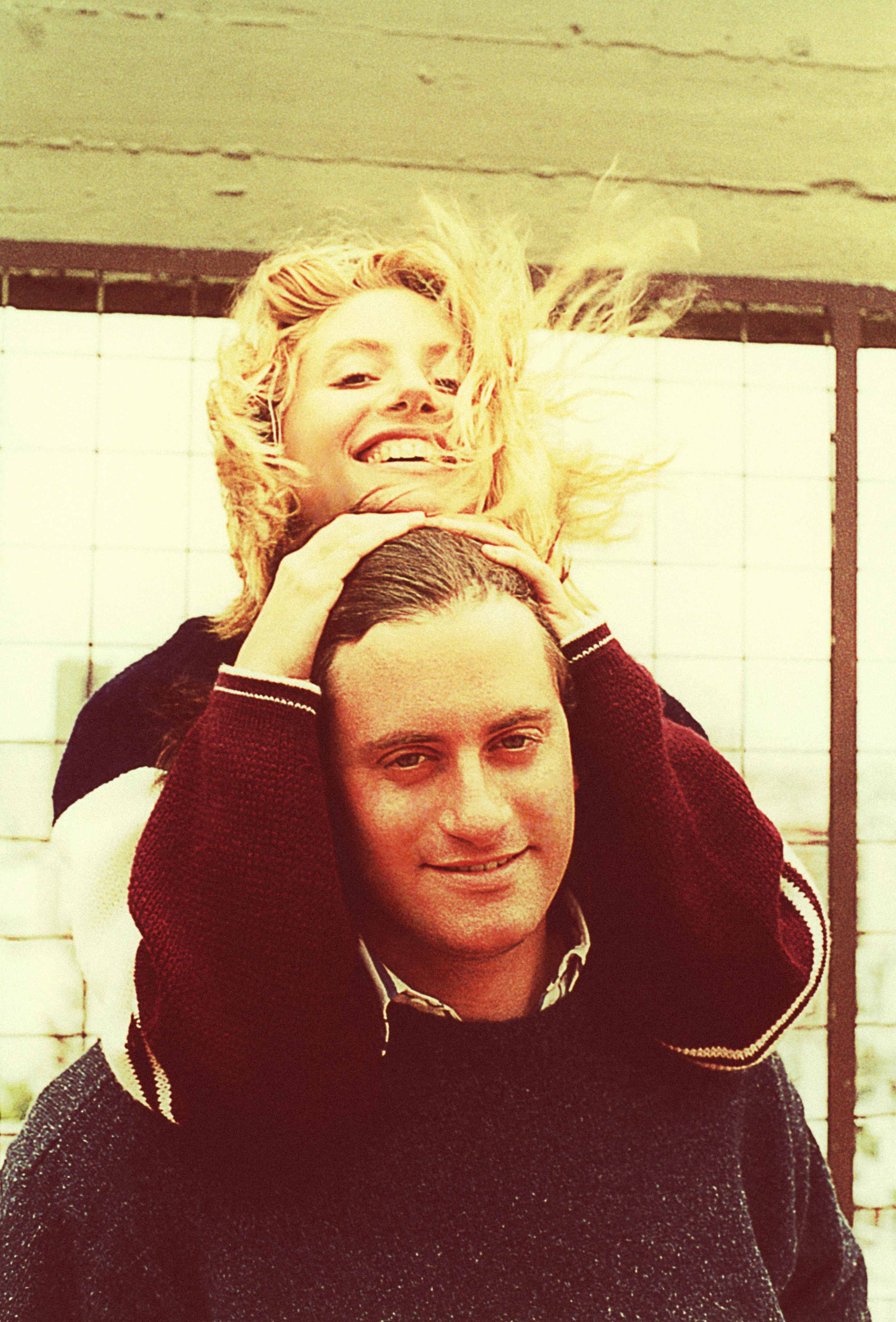 Man and woman smiling; woman playfully rests on man's shoulders, holding his head. They appear joyful and close. Casual and candid setting