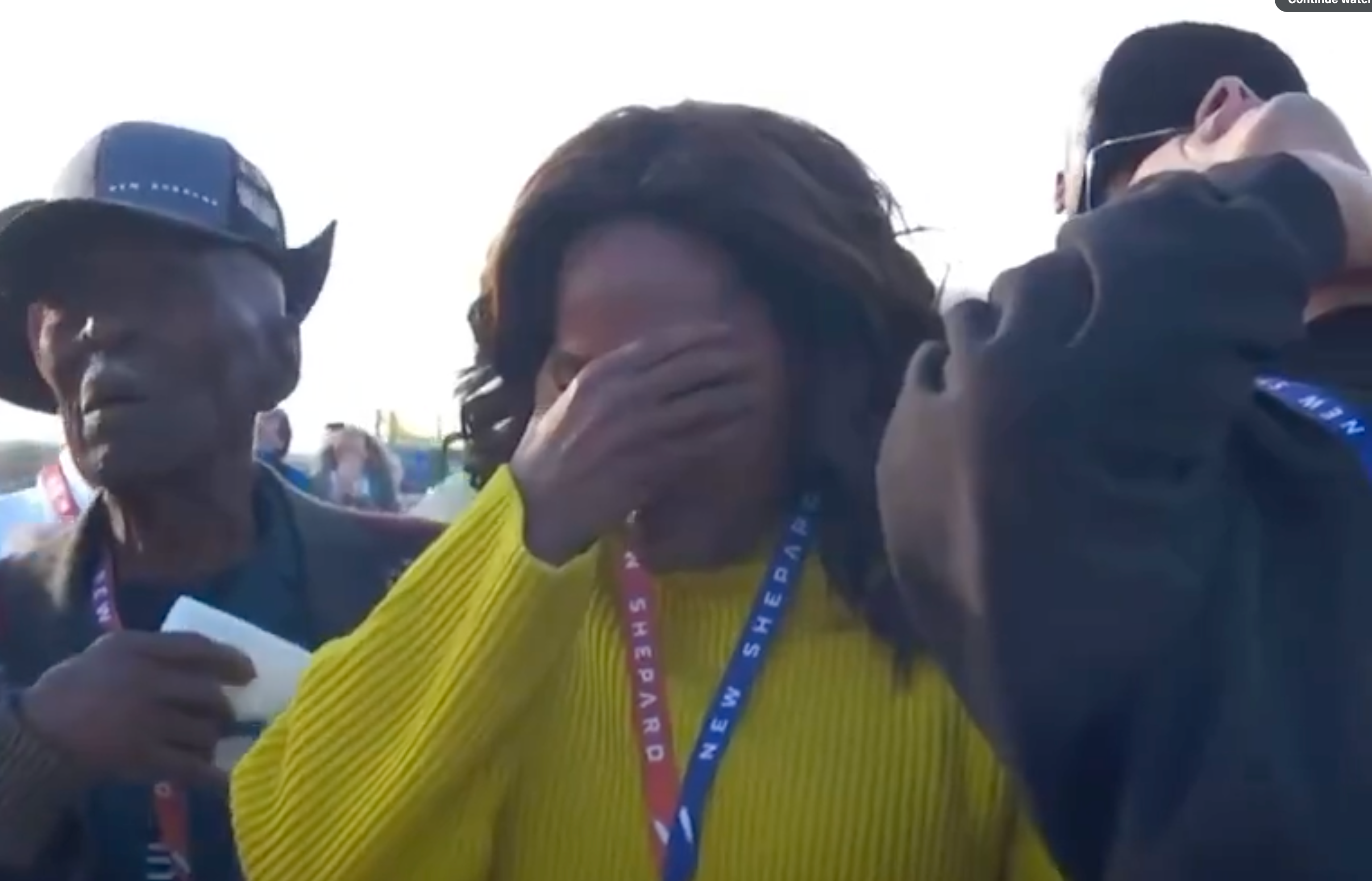 A person in a yellow sweater wipes tears, flanked by others wearing &quot;New Shepard&quot; badges at a public event
