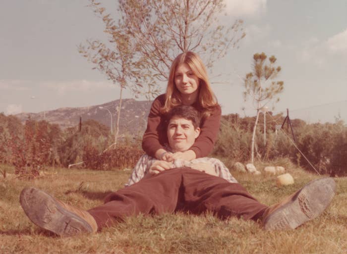 A couple is sitting on grass, with the woman behind the man, both smiling. They are embracing in a relaxed outdoor setting