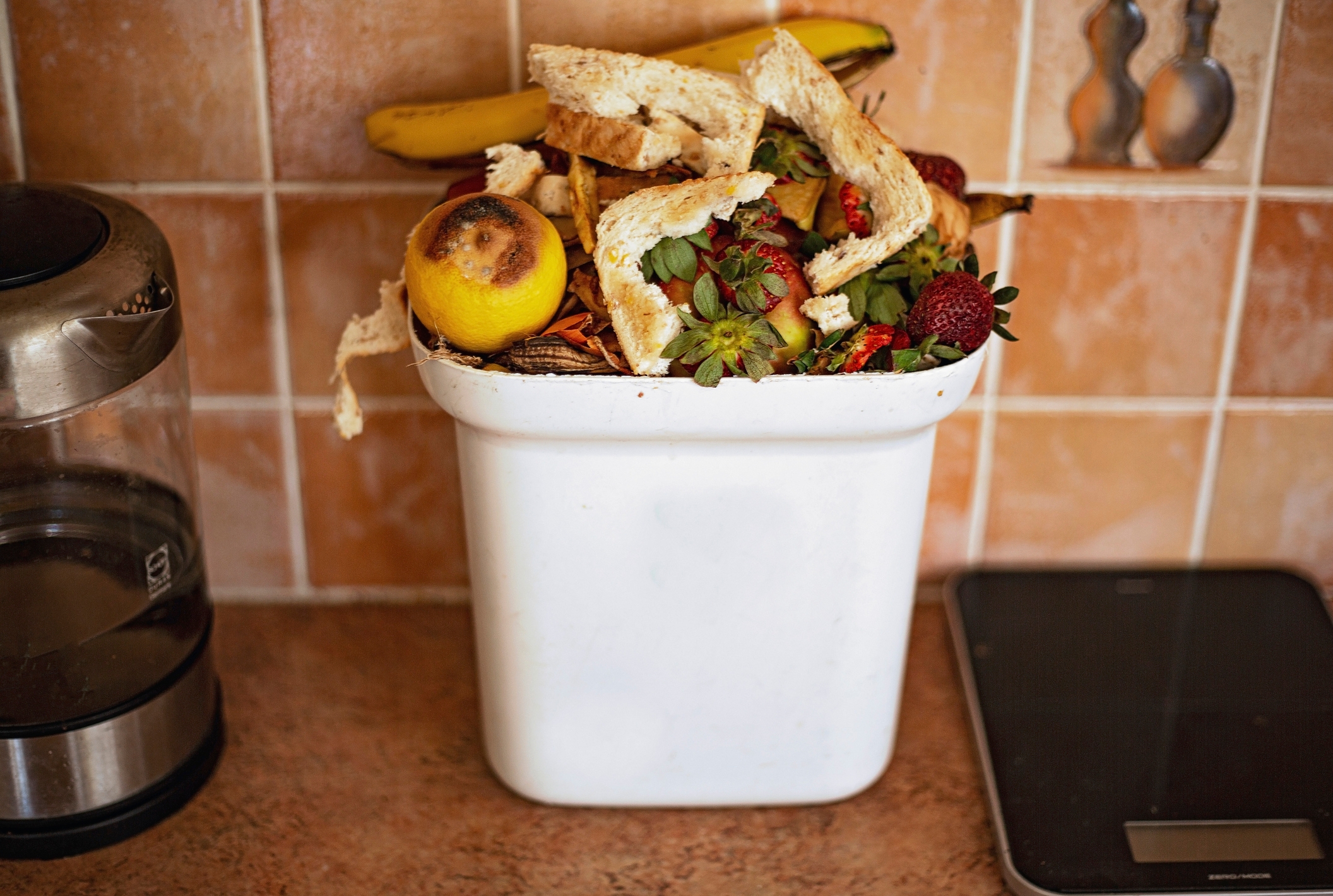 Overflowing kitchen compost bin with fruit peels, bread, and food scraps, next to a coffee maker and scale
