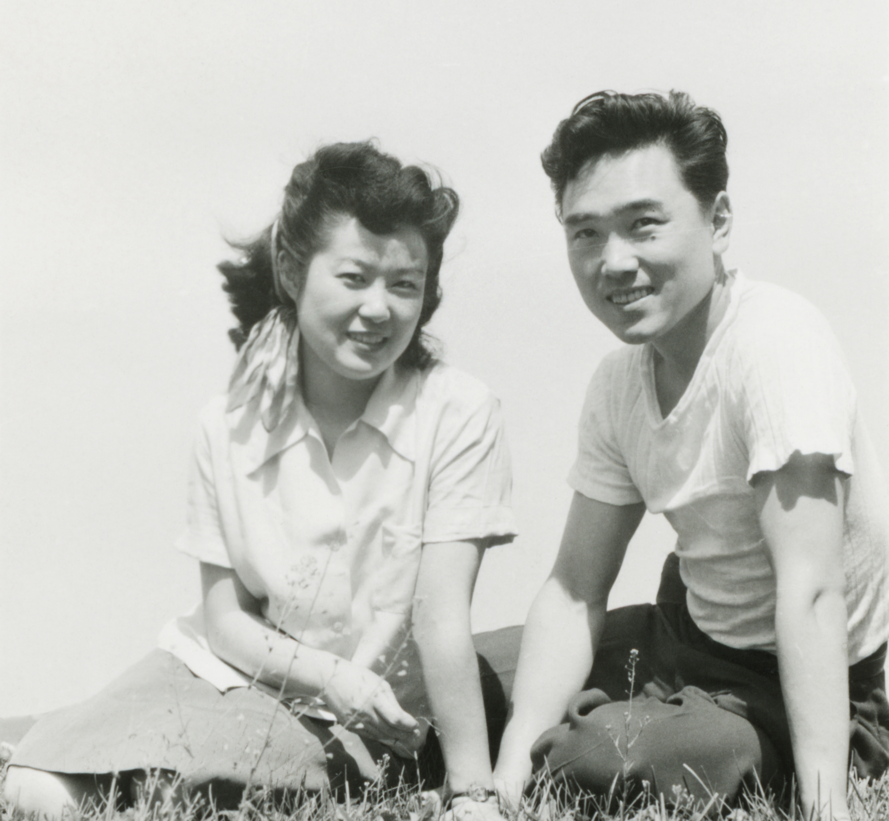 A smiling couple sits on grass, casually dressed in shirts and looking directly at the camera