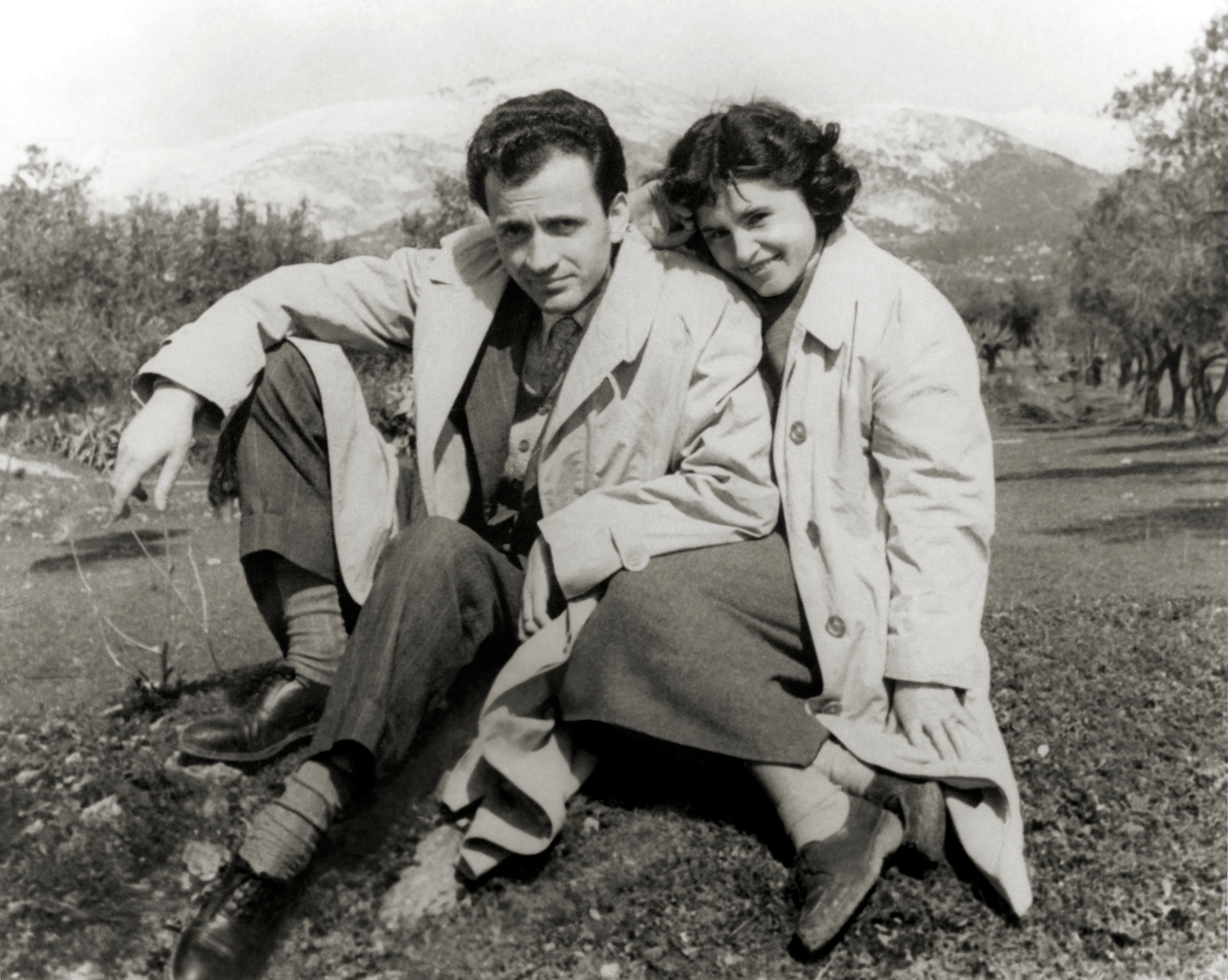 A couple in trench coats sit close together on a grassy area, smiling, with a landscape featuring distant hills in the background