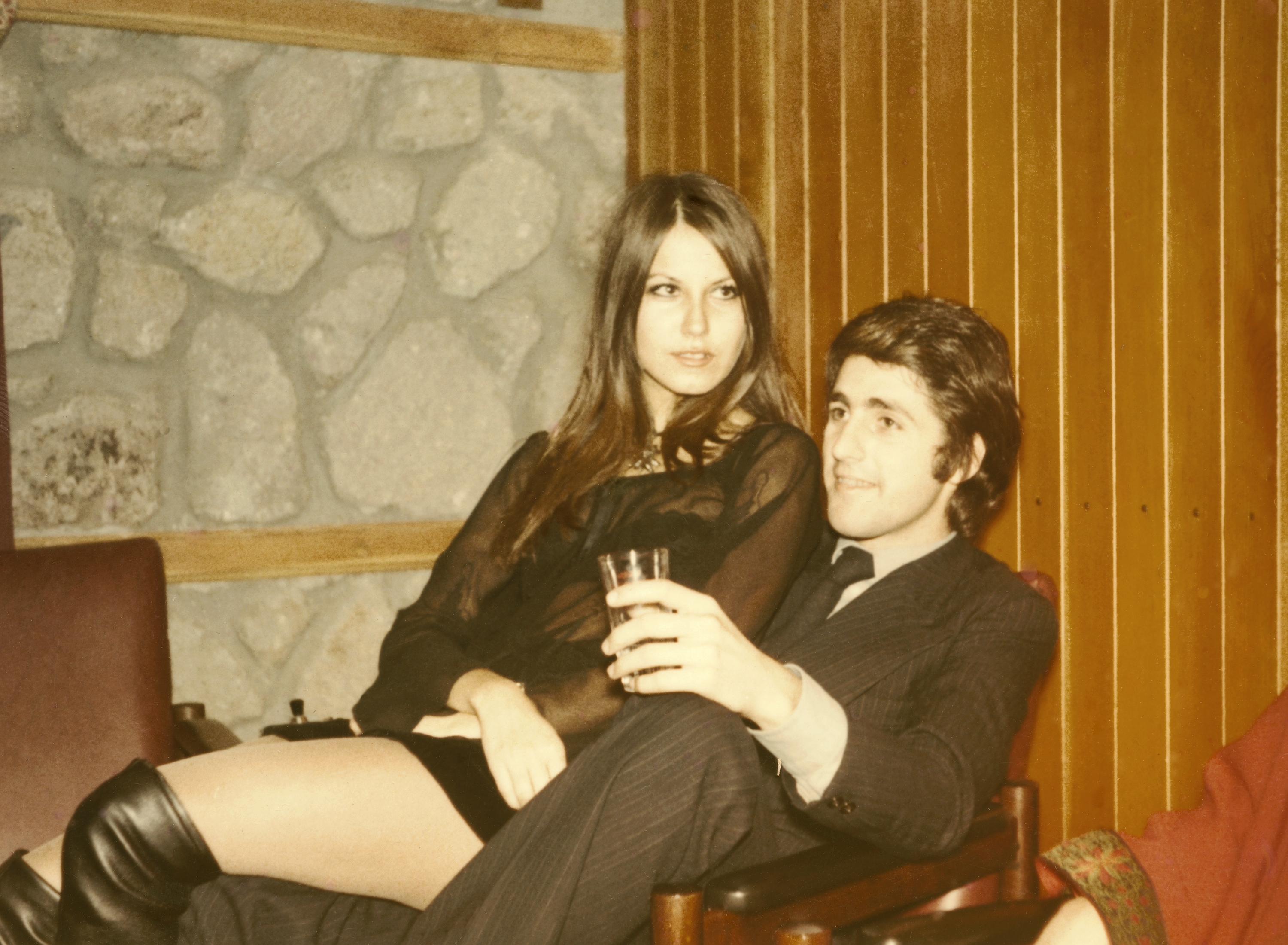 A woman in a sheer top and knee-high boots sits with a man in a suit holding a drink, both appearing relaxed in a cozy setting