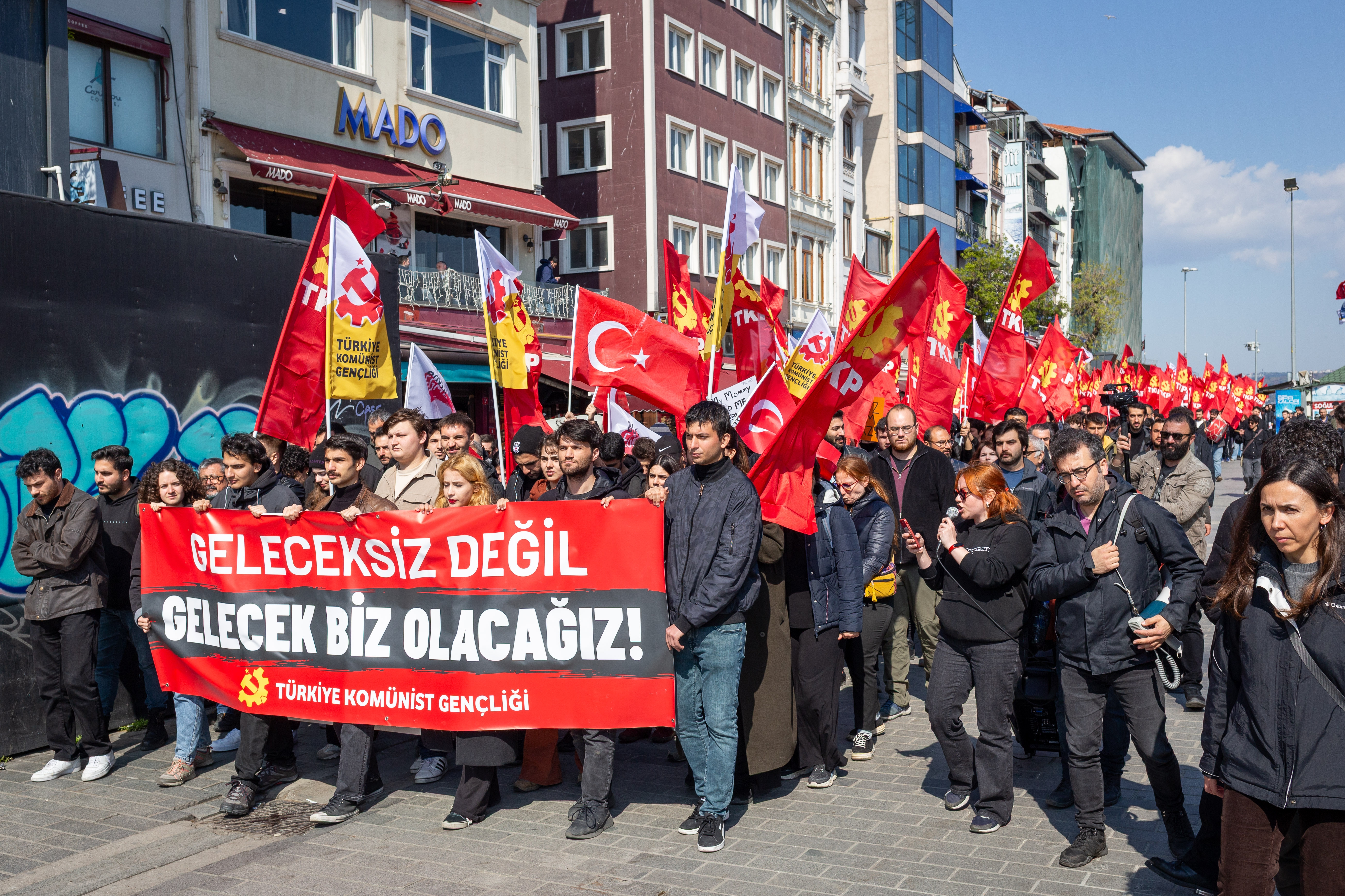 People marching in a street protest, holding red flags and a large banner with text in Turkish, surrounded by a cityscape