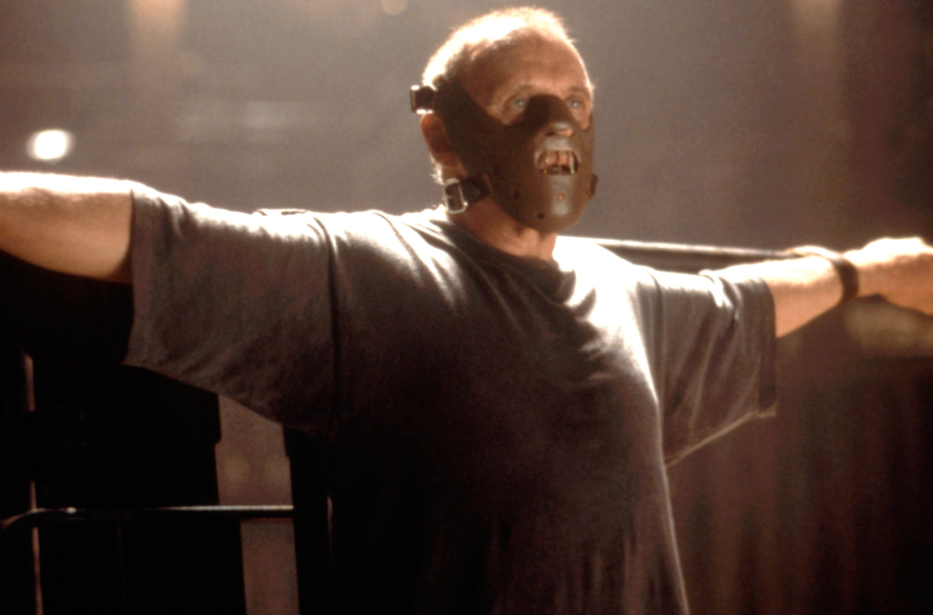 A person, restrained with arms outstretched, wears a mask with metal bars, evoking a sense of confinement