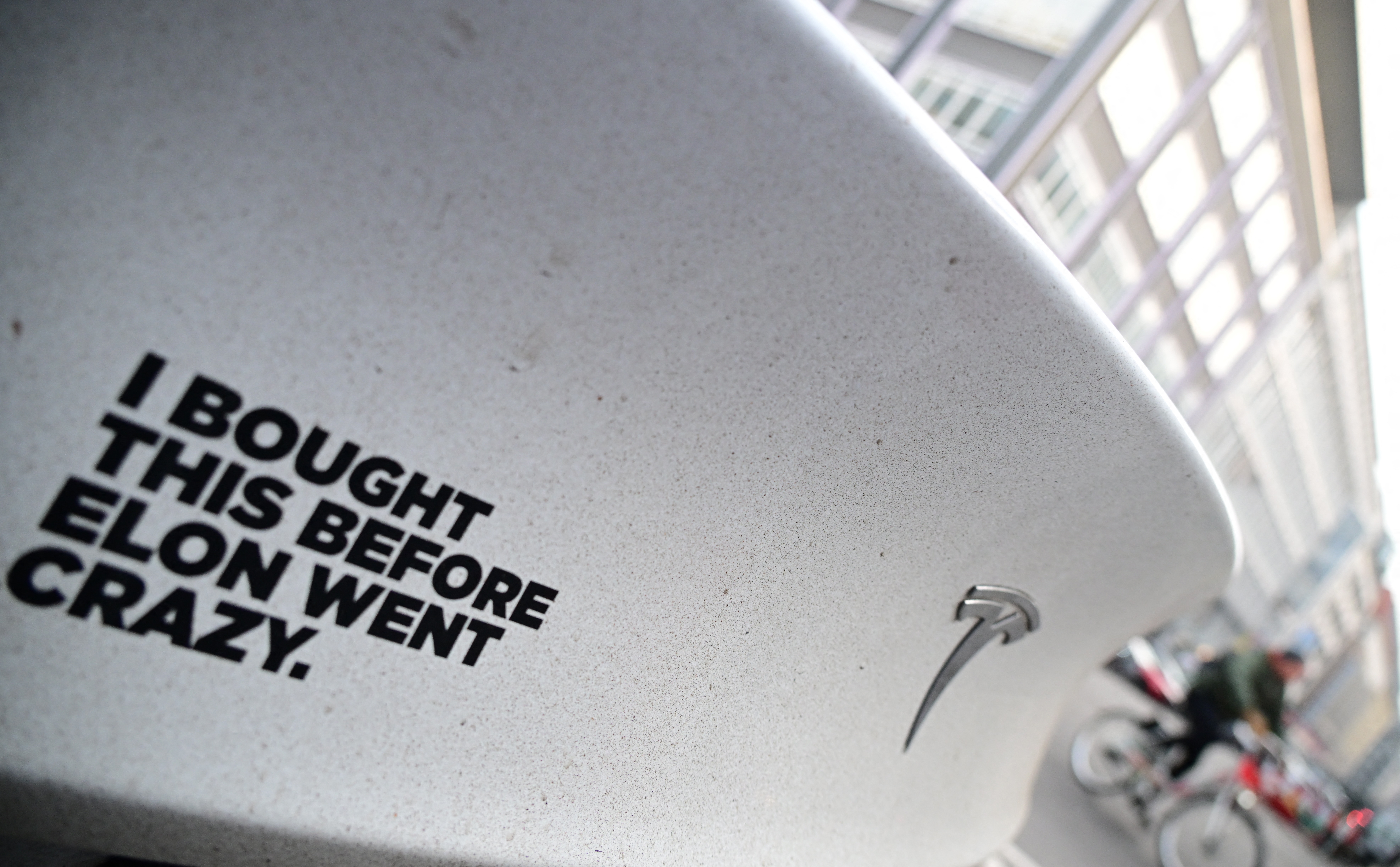 Tesla with rear sticker reading, "I bought this before Elon went crazy," seen on a city street with buildings and a cyclist in the background