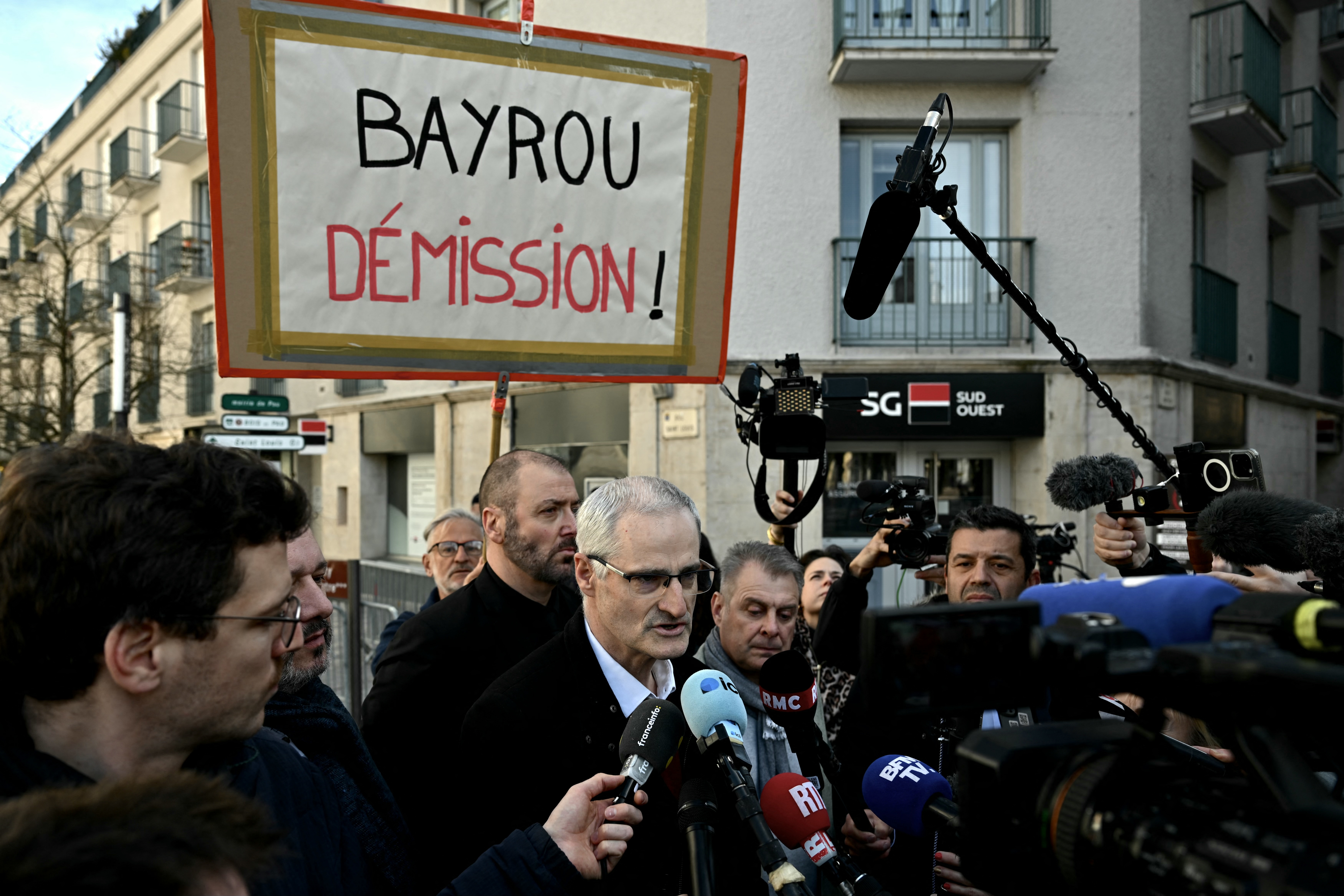 A man speaks to the media, surrounded by microphones. A protest sign reading "Bayrou Démission!" is visible above the crowd