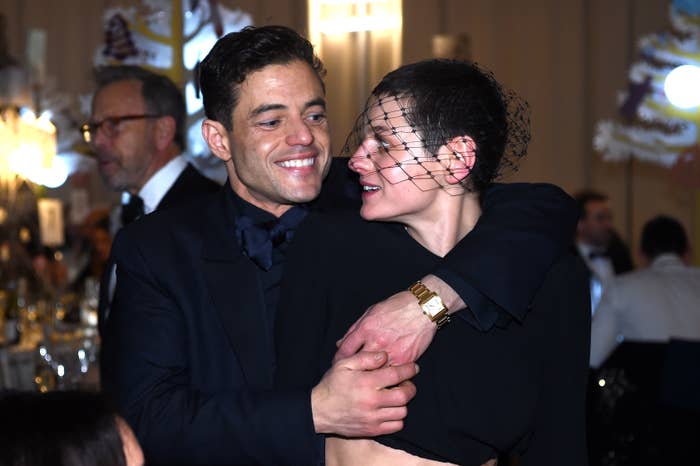 Rami Malek and Emma Corrin embrace and smile at an indoor event. The person on the right wears a black outfit and a net veil over their face