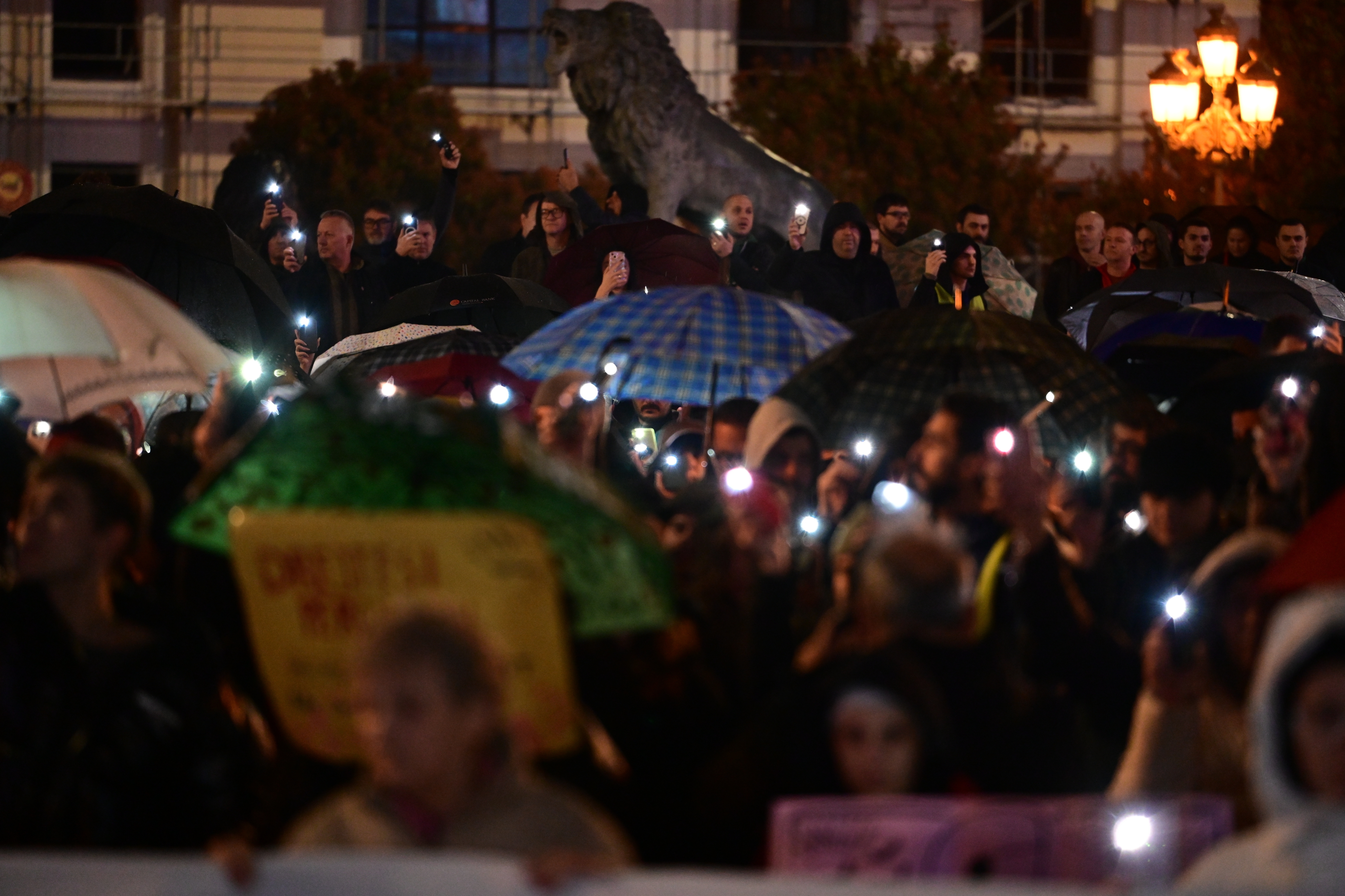 Crowd gathered at night holding umbrellas and shining phone lights, with a large statue in the background