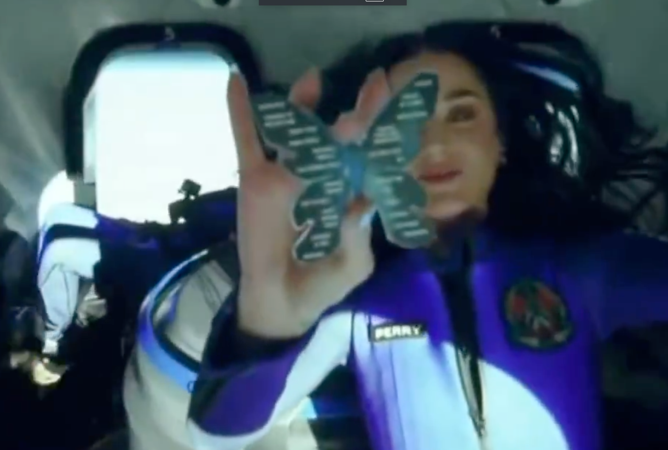 Person in a space capsule experiencing zero gravity, holding a butterfly decoration or patch, wearing a space-themed suit with a mission patch