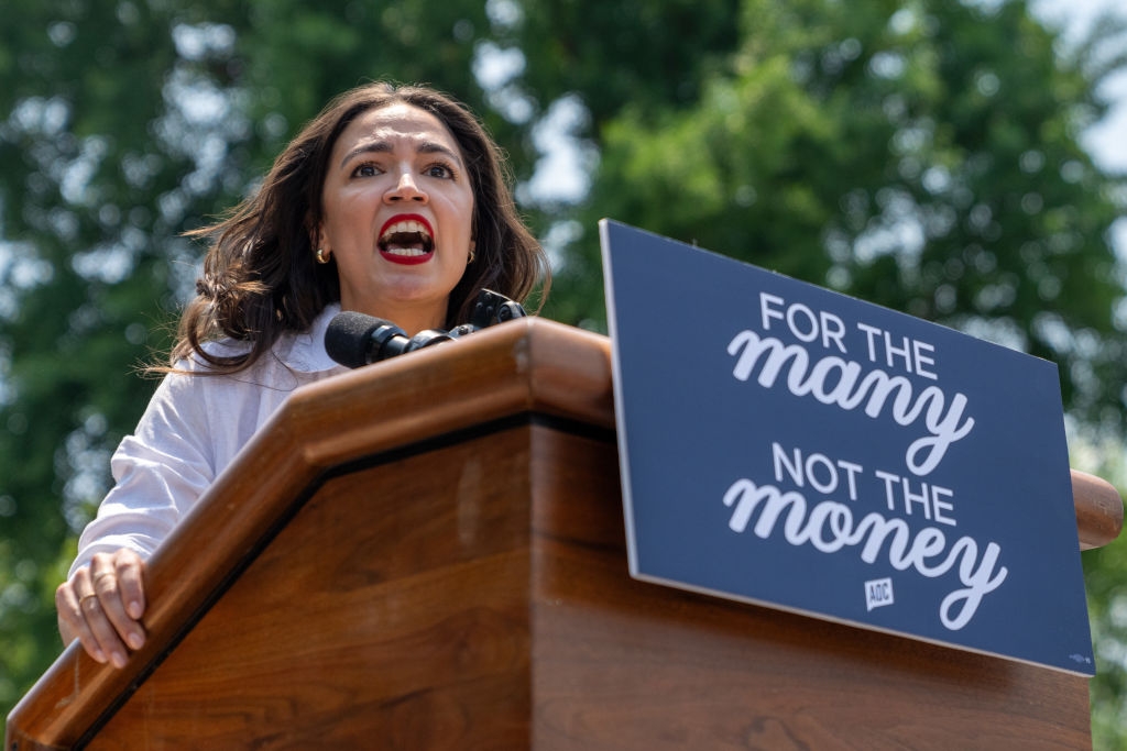 Person speaking passionately at a podium outdoors. A sign on the podium reads, "For the Many, Not the Money." Trees are visible in the background