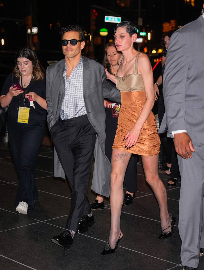 A person in a long coat with sunglasses and another in a short, strapless dress walk together at a nighttime event surrounded by people