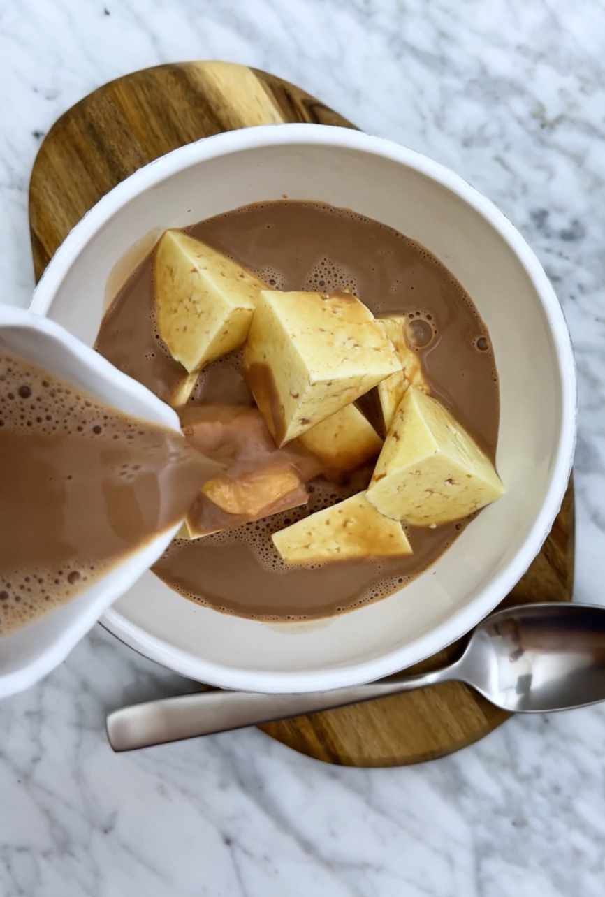 Bowl of soup with large chunks of tofu being poured with a creamy broth, set on a marble table with a wooden board and a spoon nearby