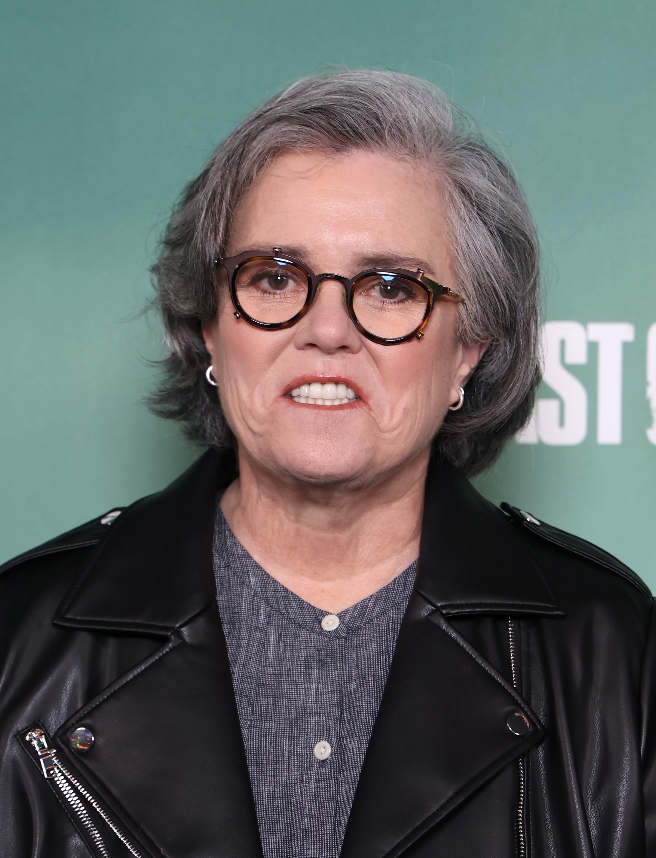 Rosie O'Donnell wearing glasses and a leather jacket, smiling slightly in front of a green backdrop