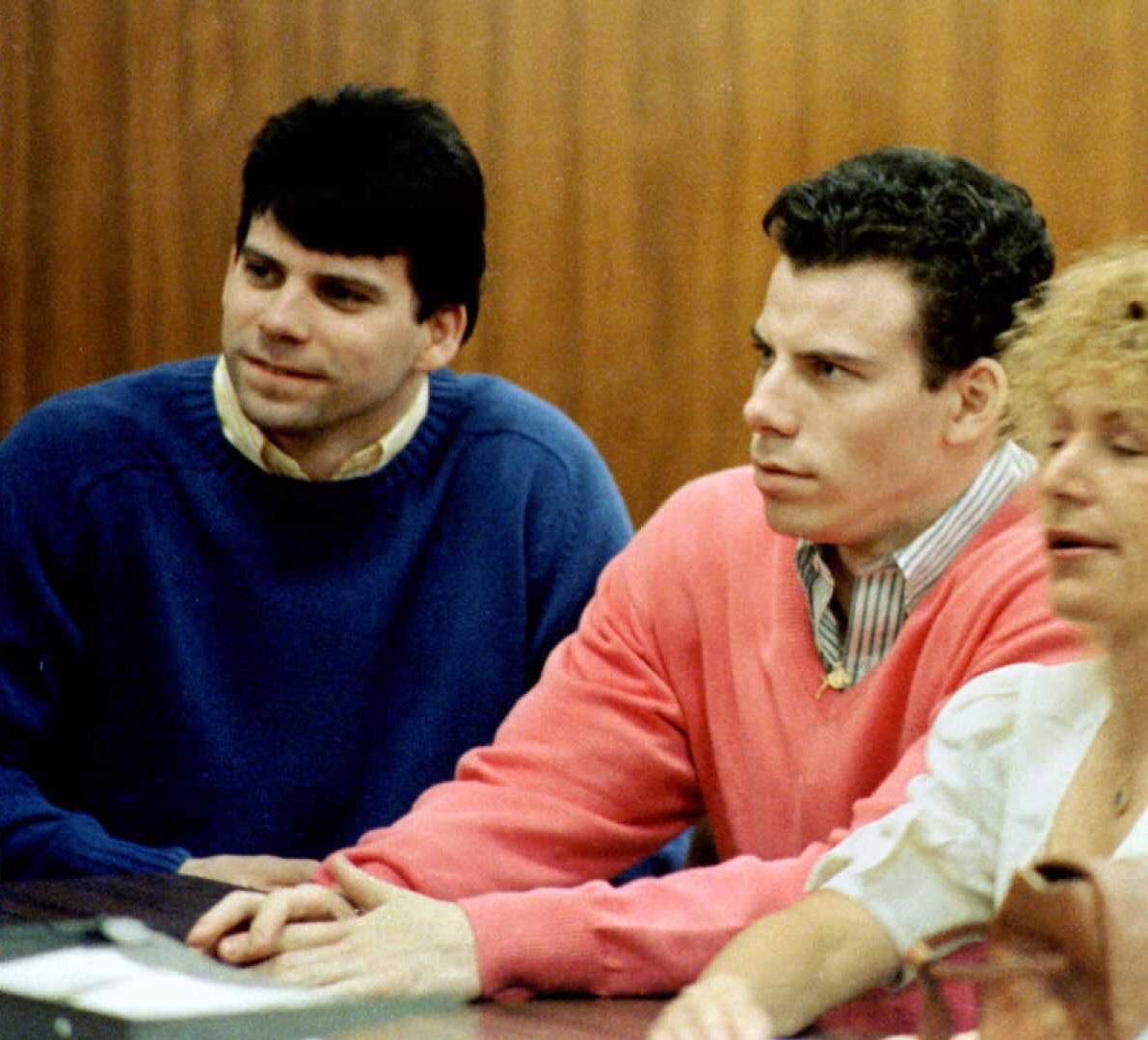 Lyle and Erik Menendez seated side by side wearing sweaters, attentively looking ahead, with another person partially visible in front
