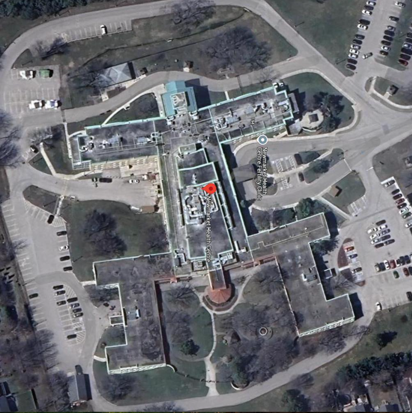 Aerial view of large building with surrounding parking and green spaces. The structure is T-shaped and details of the site are visible