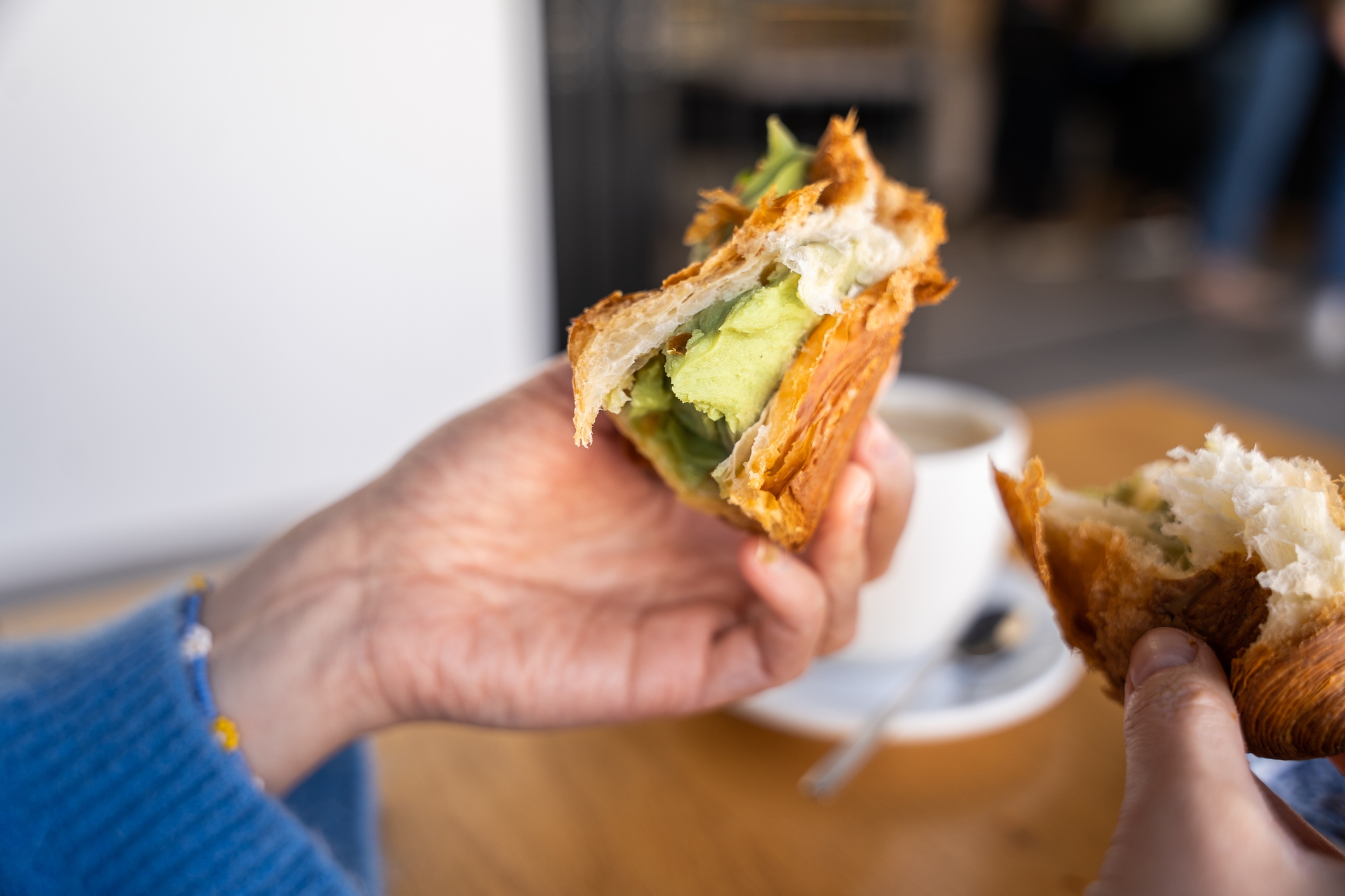 A person holds a croissant filled with avocado. Another hand holds a piece of croissant. A cup on a saucer is in the background