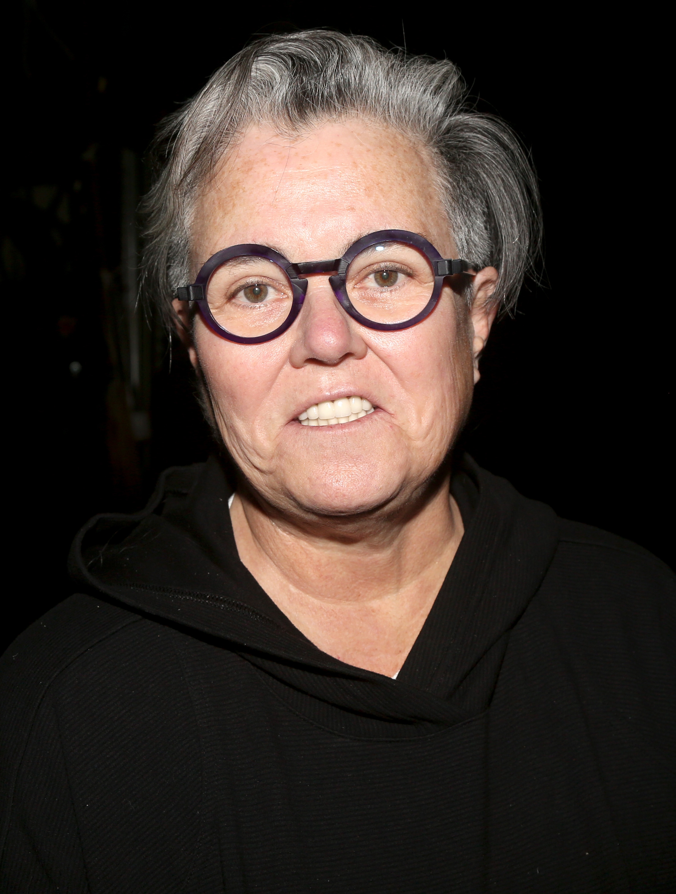 Rosie O'Donnell wearing round glasses and a dark hoodie, with short gray hair, against a dark background