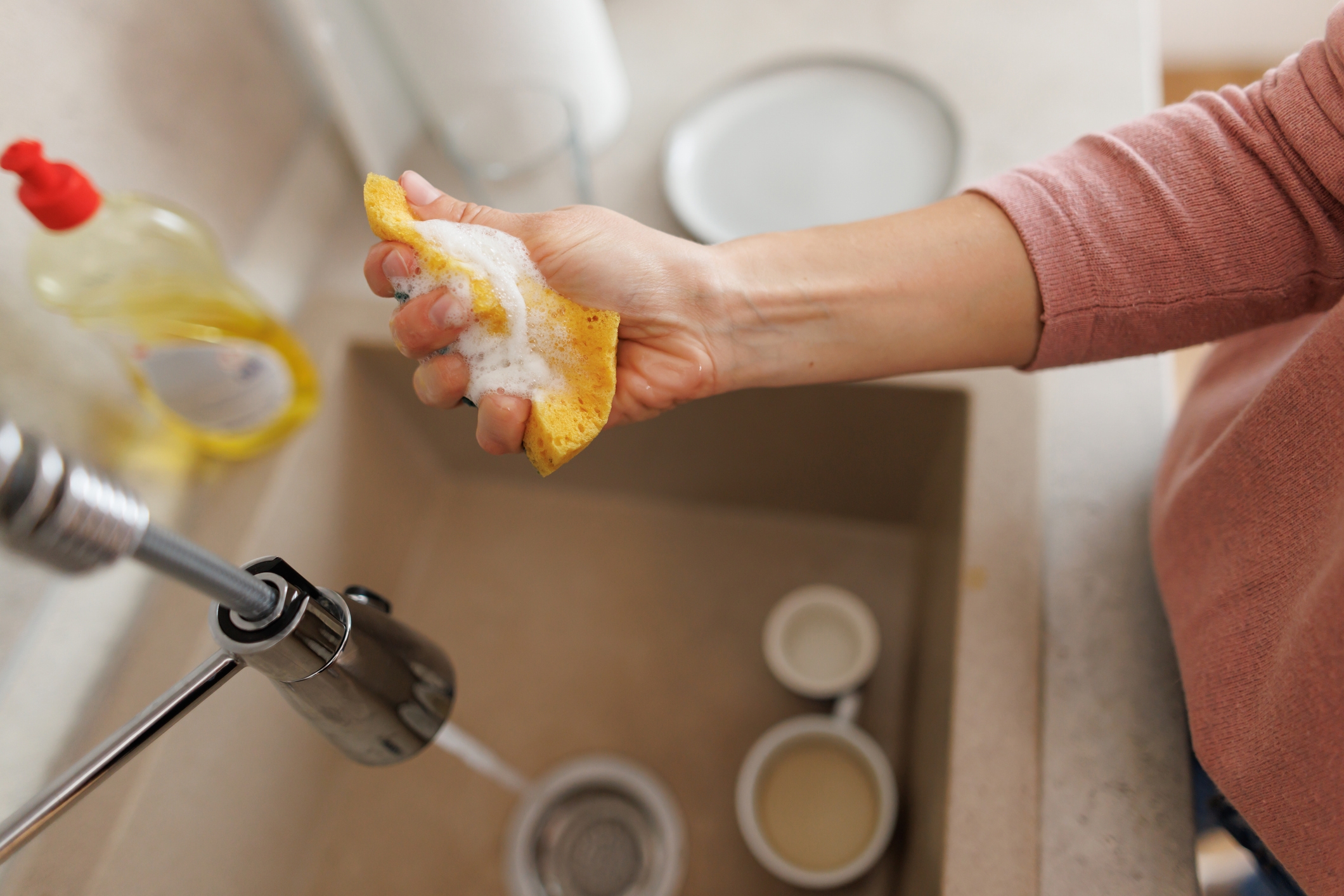 A person cleaning a kitchen sink with a soapy sponge, water running from the faucet. Dish soap and containers are visible on the countertop