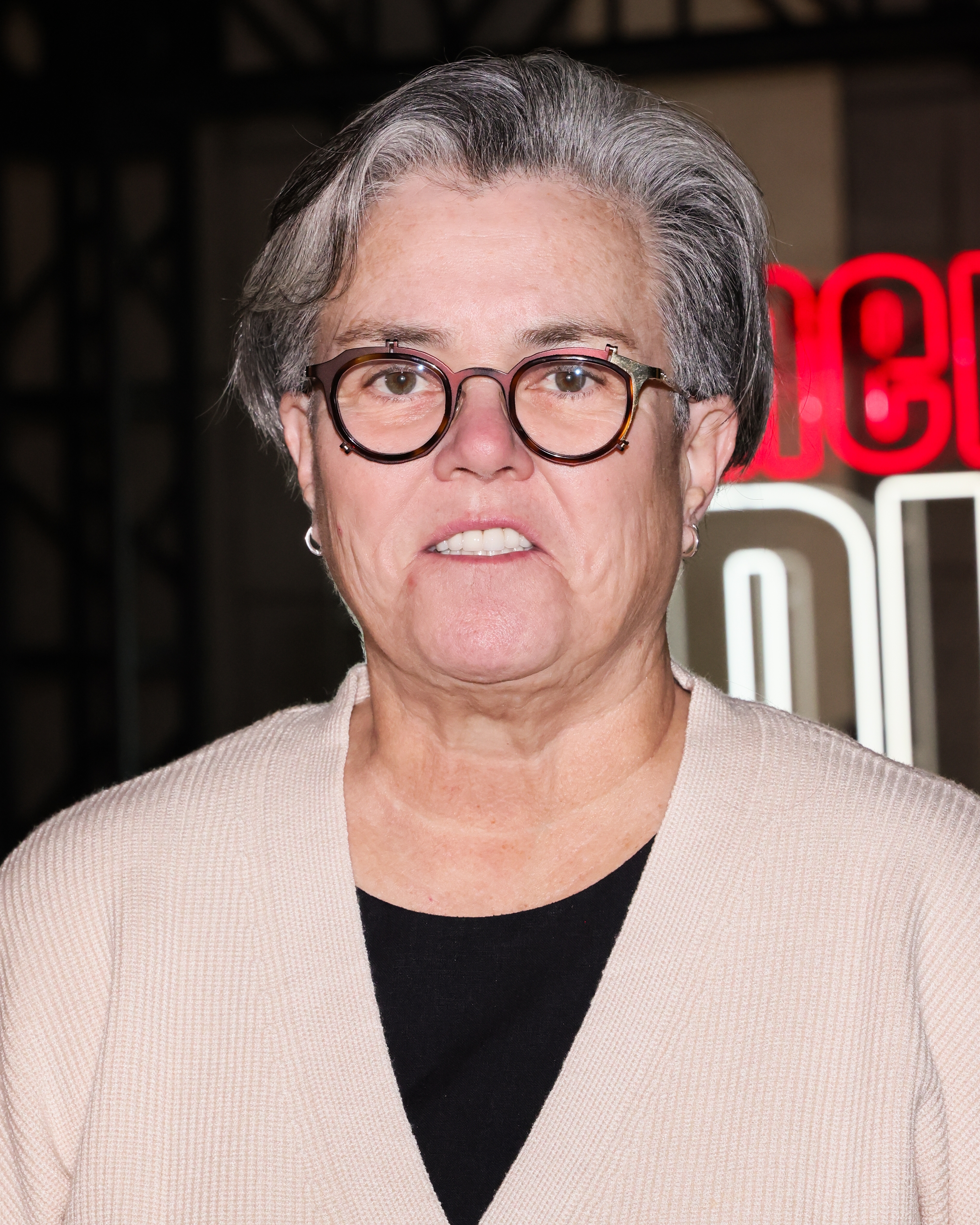 Rosie O'Donnell wearing glasses and a light cardigan over a dark top, in front of a neon sign backdrop