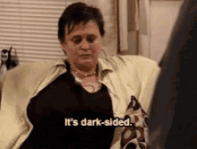 Person sitting, gesturing while saying, "It's dark-sided," in a casual indoor setting