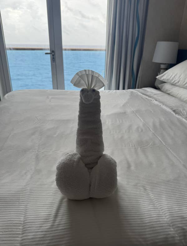 A towel elephant sculpture with a fan-shaped head sits on a neatly made bed in a room with an ocean view through the window