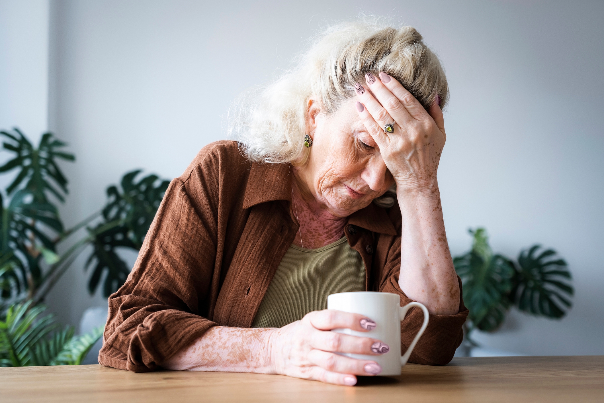 Eldre kvinne sitter ved et bord og holder hodet med den ene hånden og ser gjennomtenkt ut mens han holder et krus i den andre