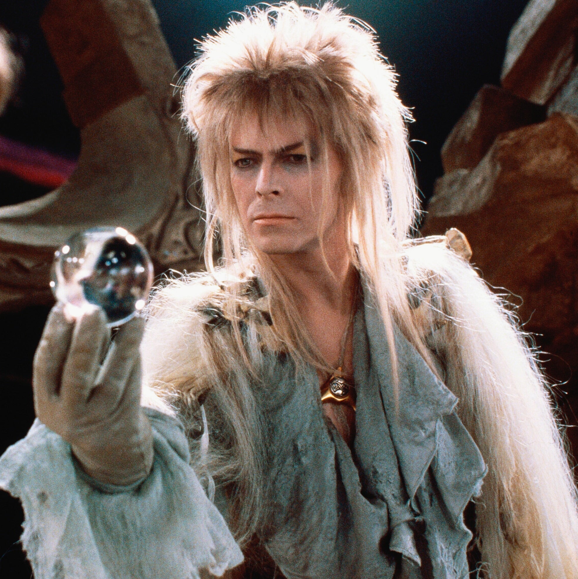 A mystical character with wild hair holds a crystal orb, gazing intensely at a focused young girl, hinting at a fantasy movie scene