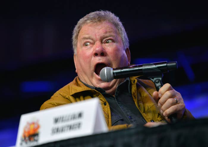 A surprised man holds a microphone at a panel, wearing a casual jacket, with an expression of shock or excitement. Nameplate visible but not readable