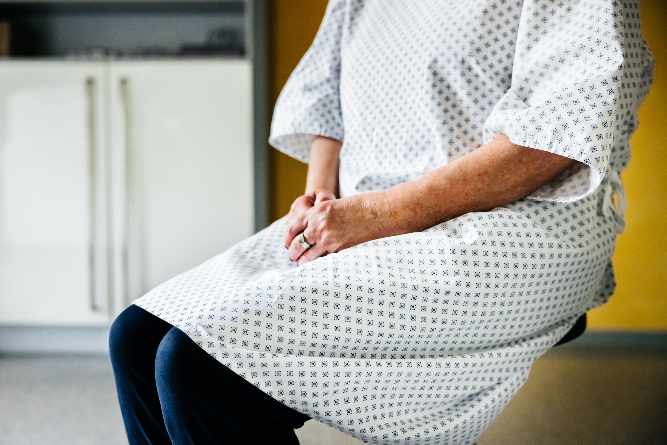 Person seated wearing a hospital gown, hands resting on lap, sitting in a medical setting with a cabinet visible in the background