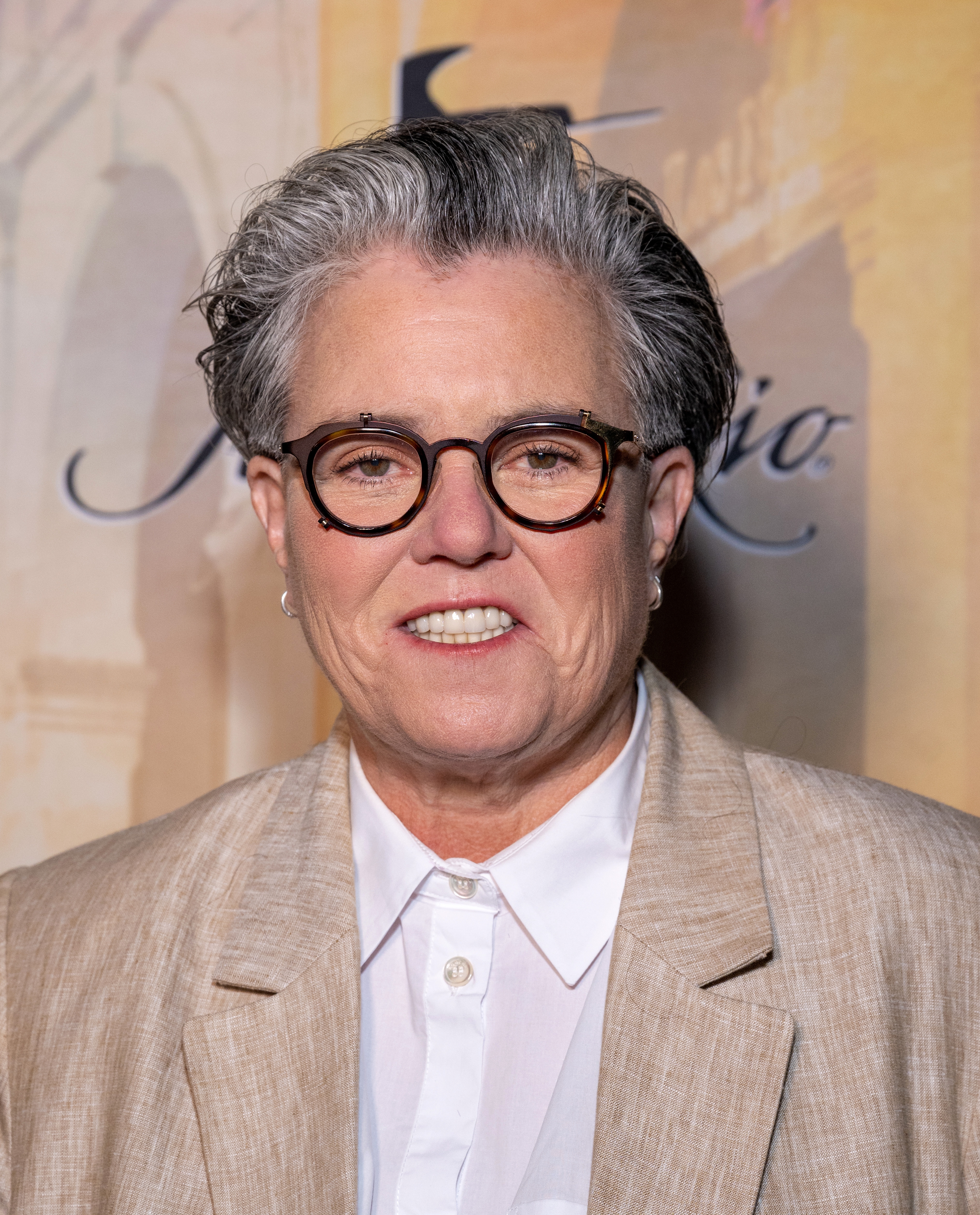 Rosie O'Donnell in a light-colored suit and white shirt, wearing glasses, stands in front of a backdrop with indistinct text