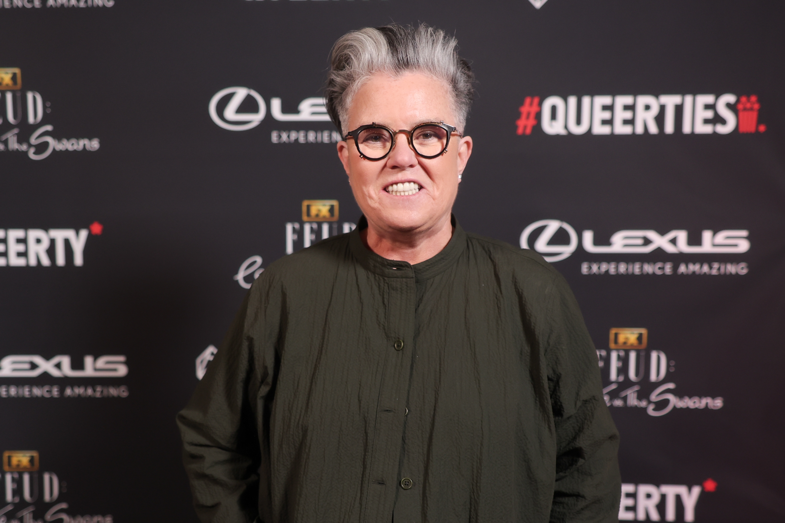 Rosie O'Donnell with short hair and glasses on a red carpet wearing a pleated dress shirt, posing in front of a backdrop with event branding