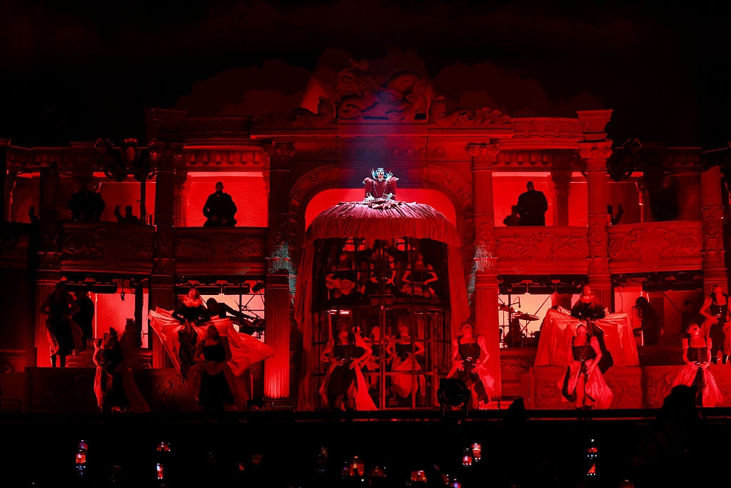 A performer in a dramatic feathered costume is elevated above a stage with dancers. The scene is set in an opulent, theater-like environment