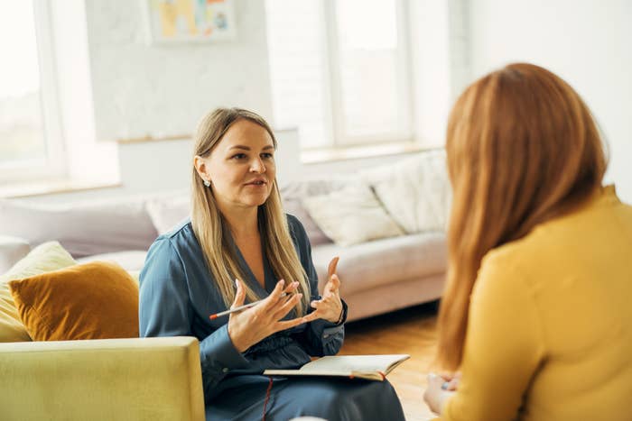 Two people sit on a couch, engaged in a conversation. One takes notes while the other gestures expressively