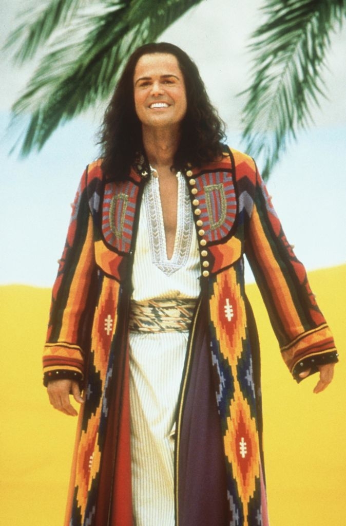 Person in colorful, patterned robe, posing outdoors, smiling. The robe features geometric designs and intricate details