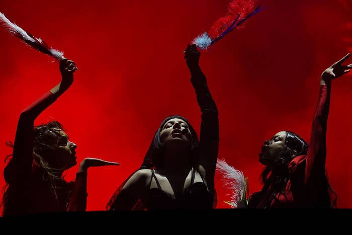 Three performers on stage strike dramatic poses holding feathers against a bold red backdrop during a show