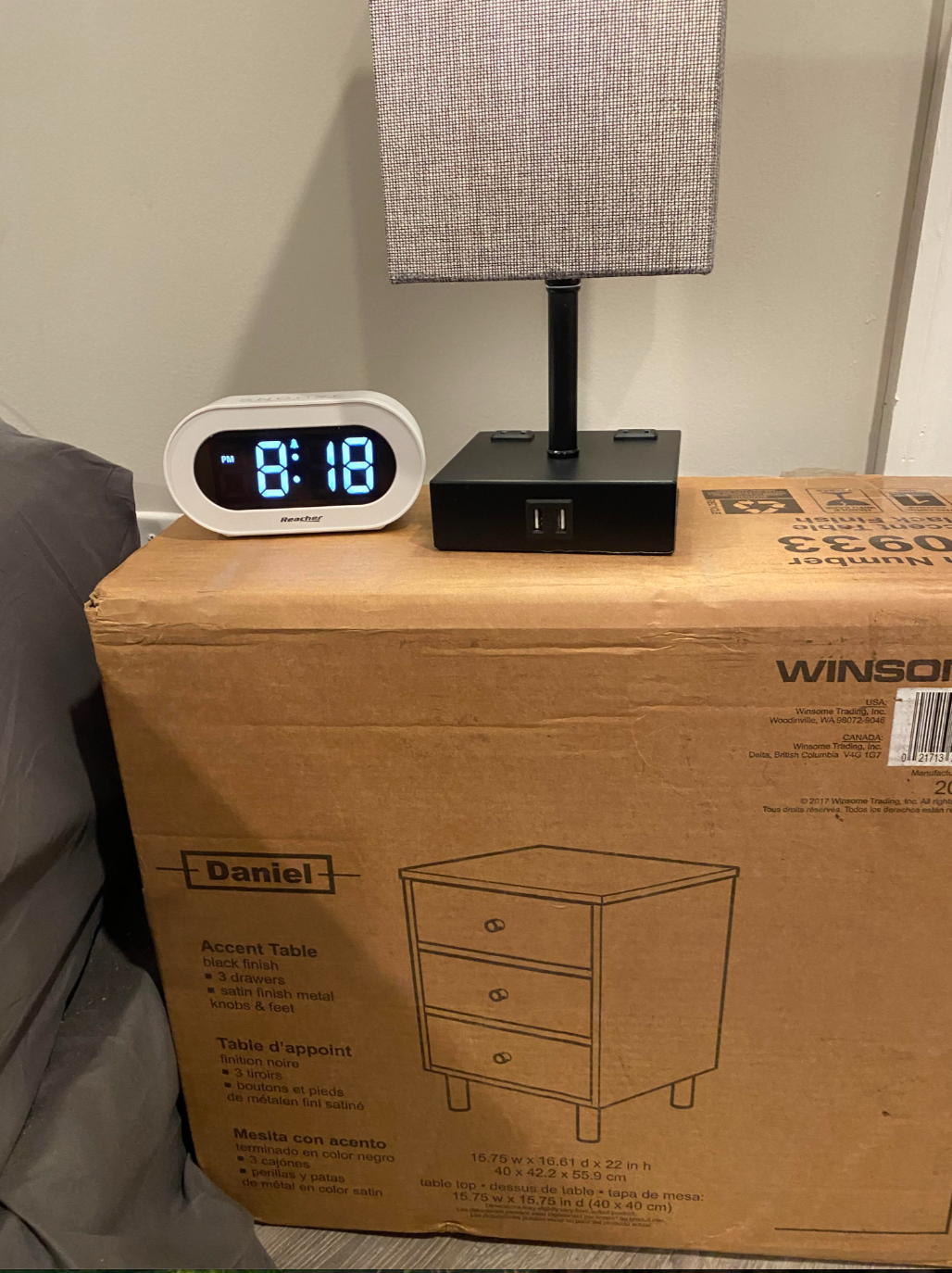 Digital clock reading 8:18 and a lamp sit atop an unopened box labeled "Accent Table." Drawer illustration on box suggests furniture style