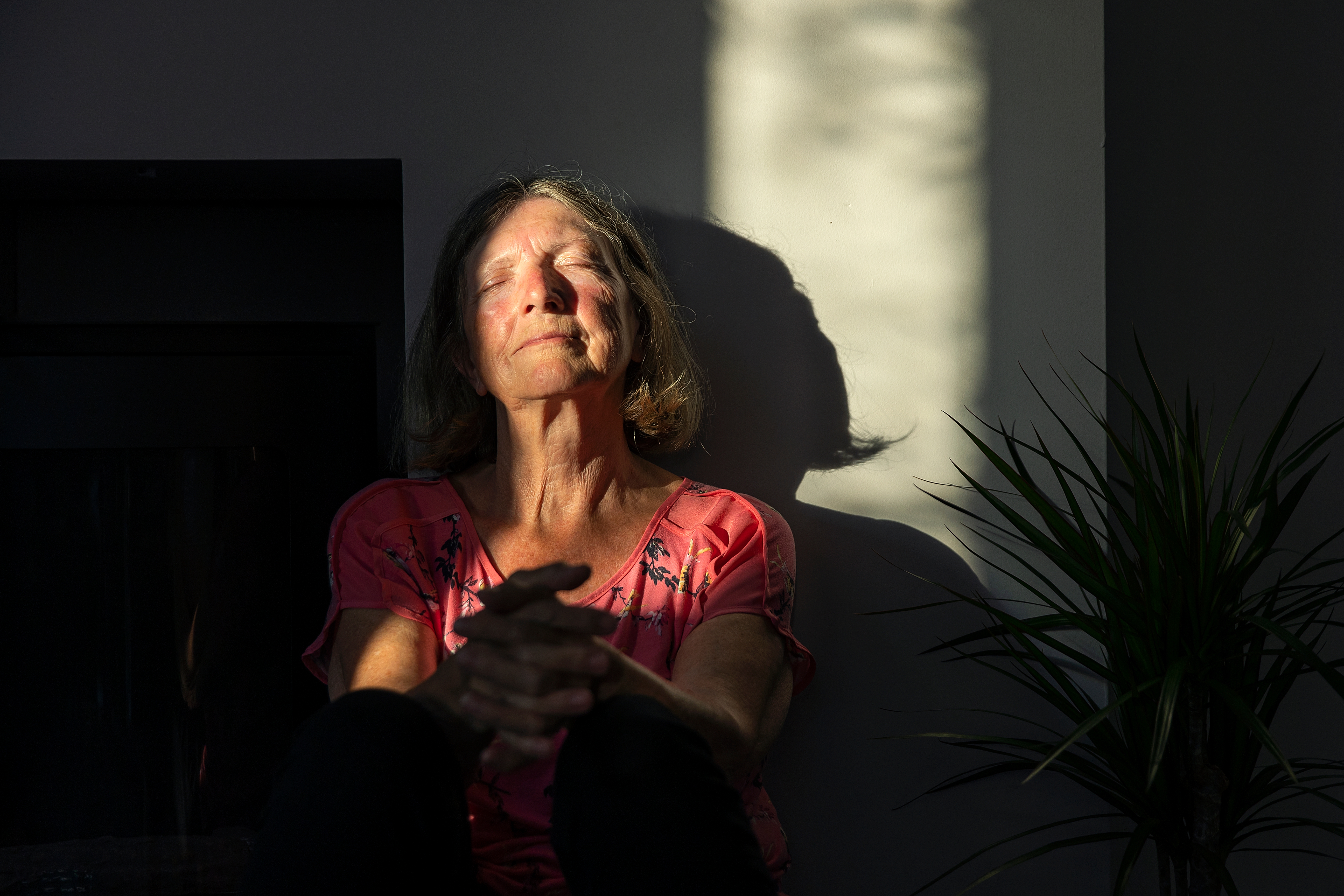 En person sitter fredelig med lukkede øyne, badet i sollys mot en vegg, ved siden av en plante