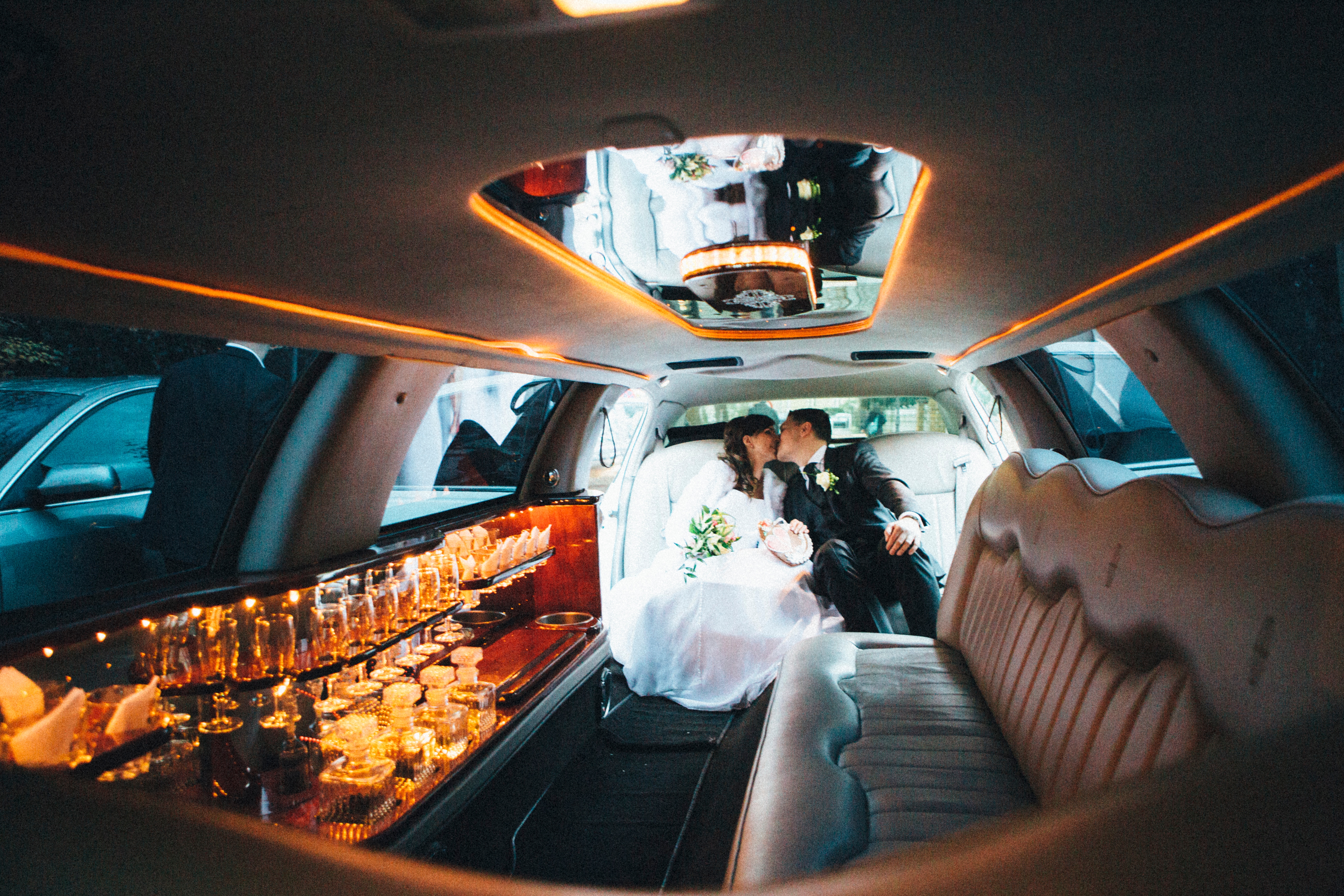 Couple in formal attire share a kiss inside a luxurious limousine, surrounded by an elegant interior and a minibar, suggesting a celebratory event