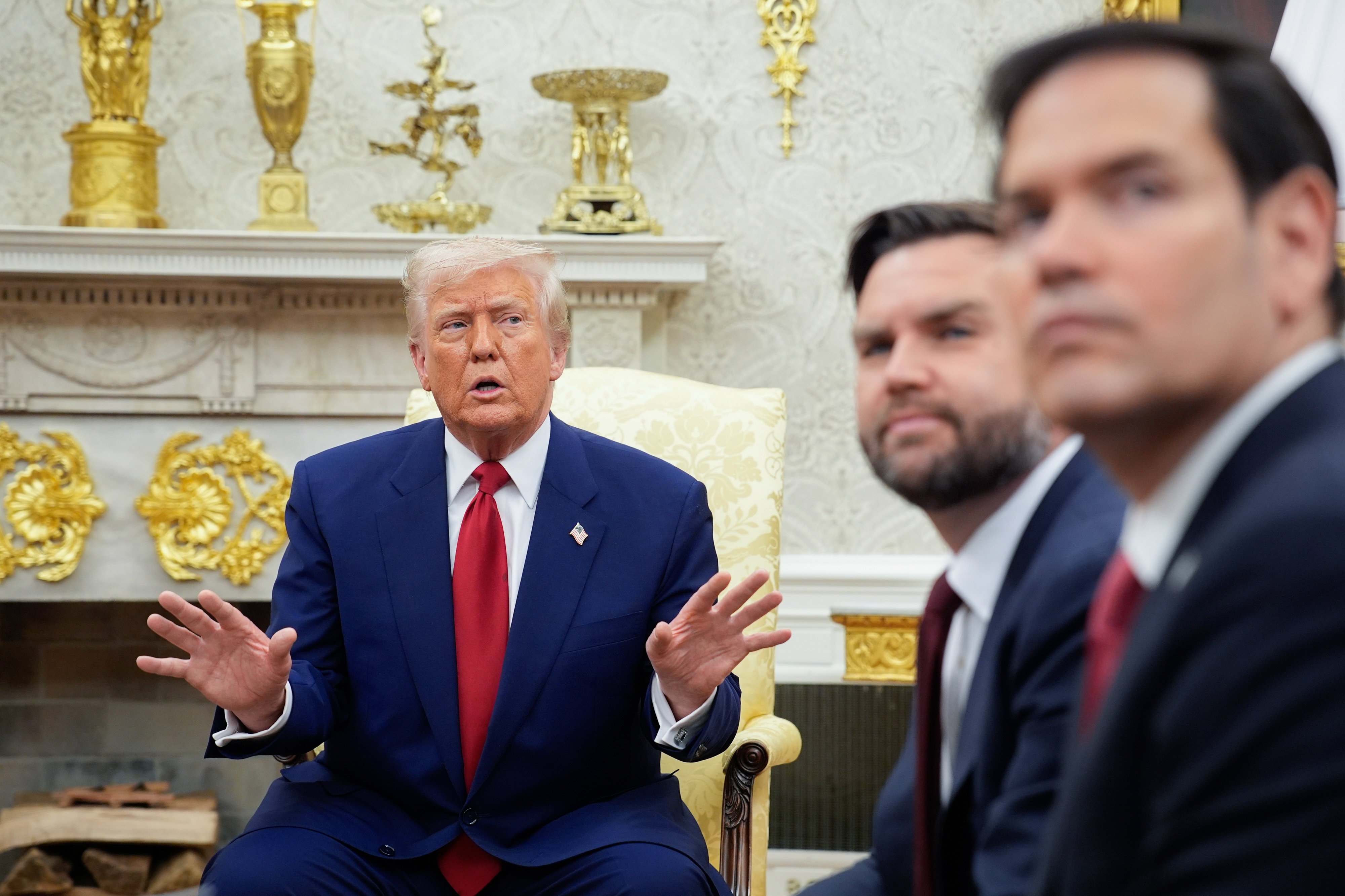 Three men in suits, sitting in a formal room. The man in the center gestures with open hands, engaged in conversation
