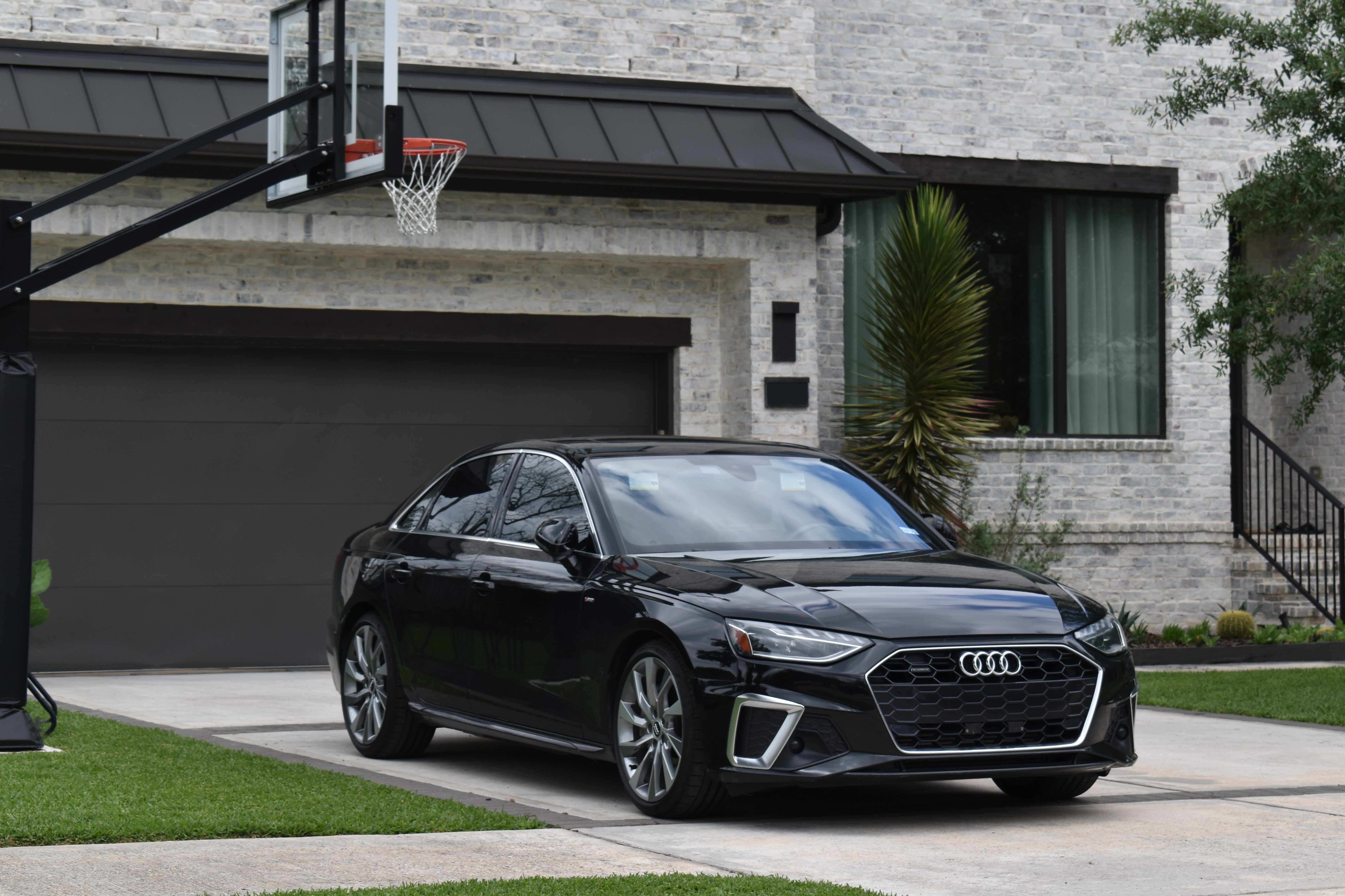 An Audi sedan is parked in a residential driveway next to a basketball hoop, in front of a modern brick house