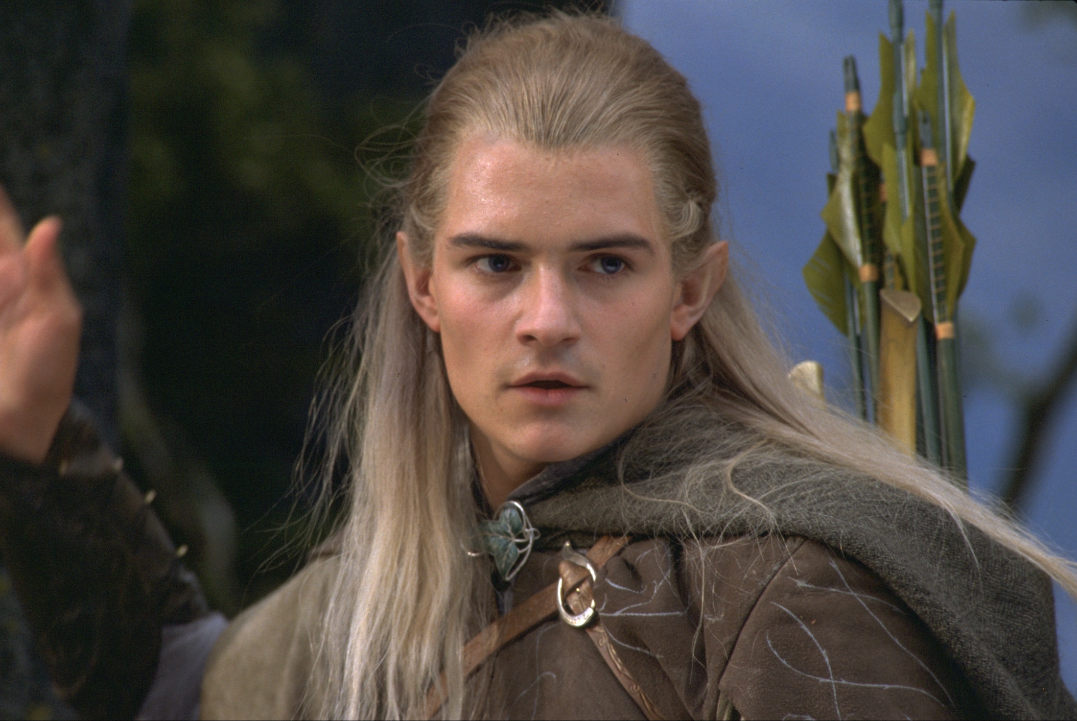 Person in fantasy costume with long hair and cloak, holding a bow and quiver of arrows, looking intently off-camera