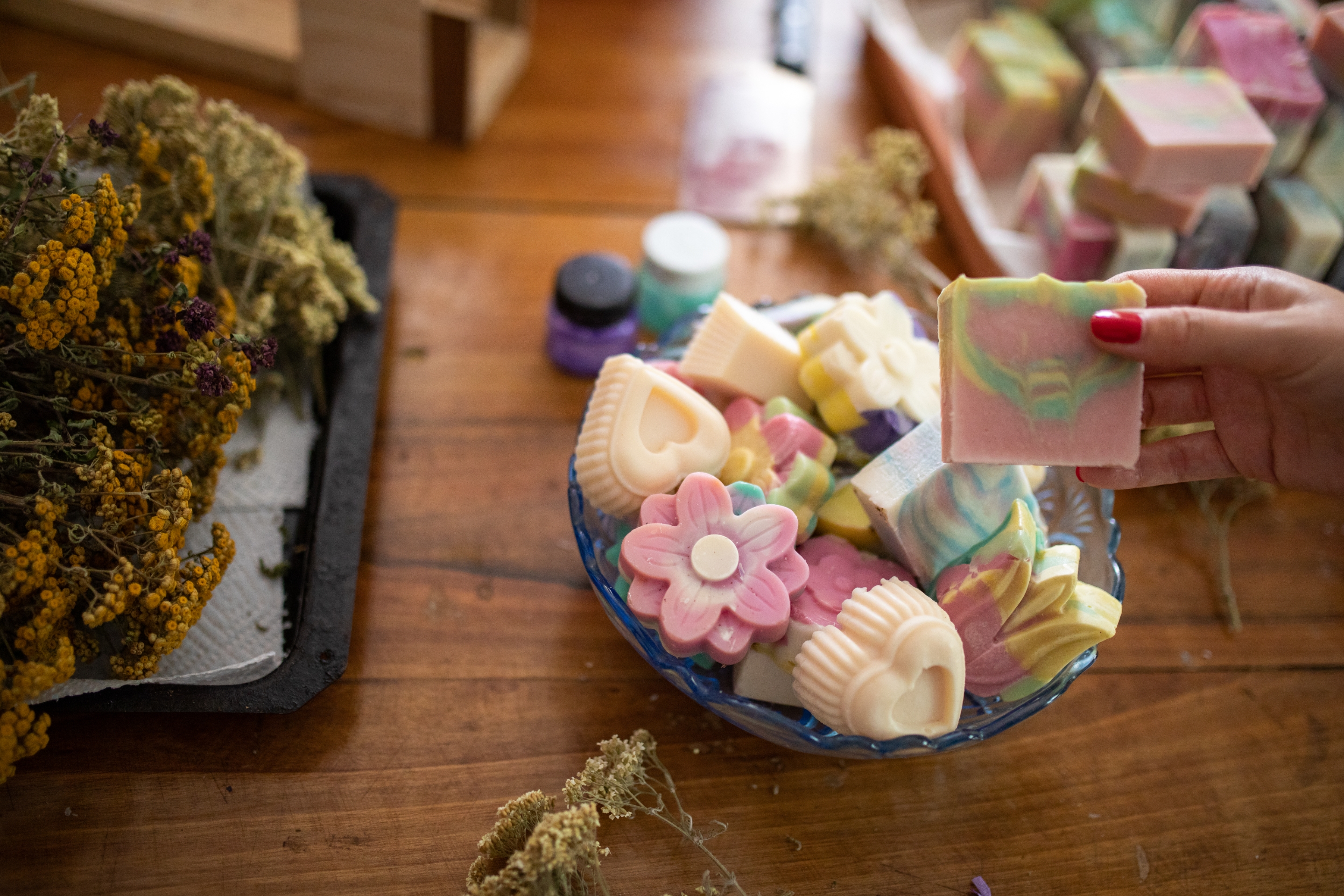 Hand holding colorful handmade soaps in various shapes near dried flowers on a wooden table, highlighting artisanal or small business work