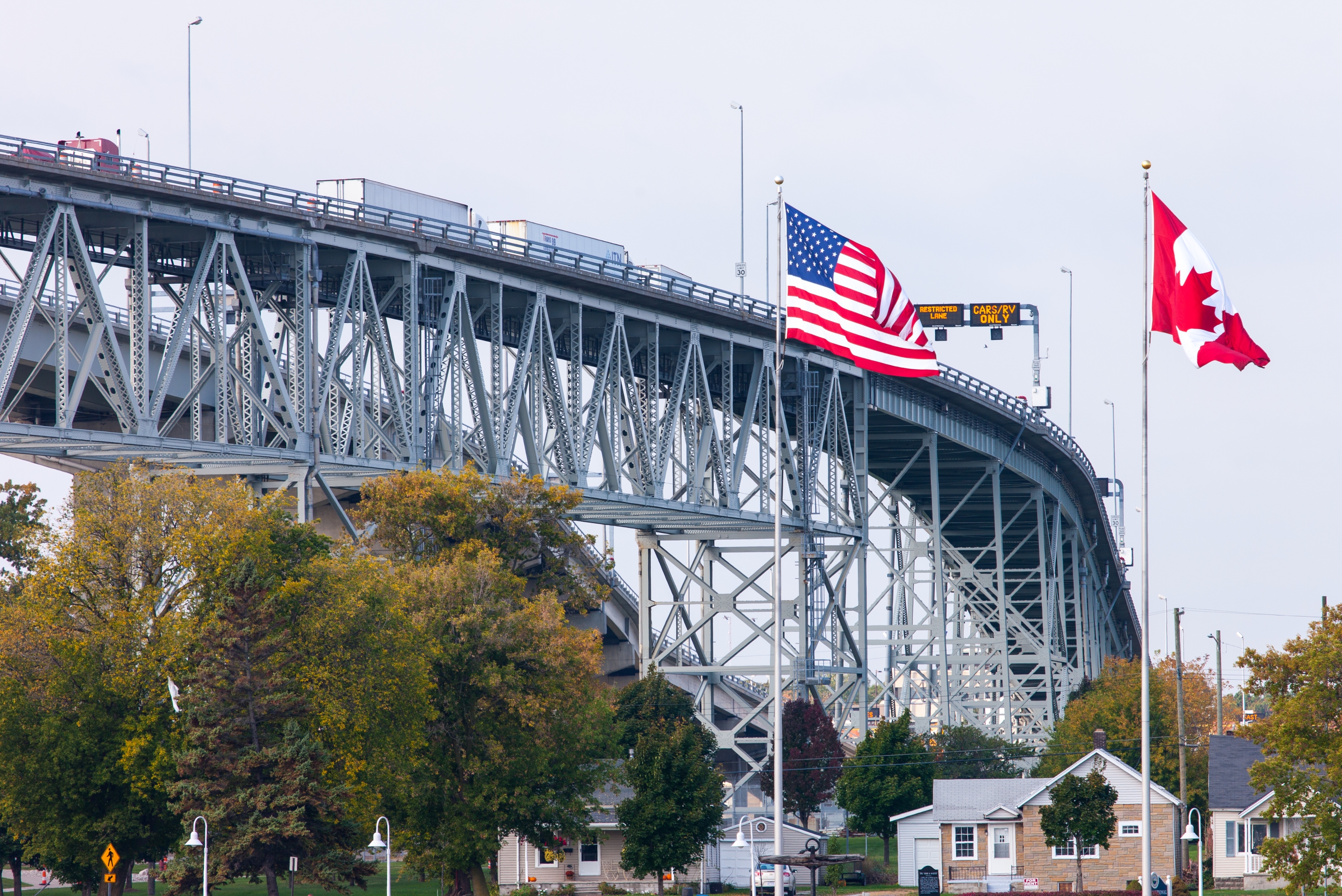 Two flags, American and Canadian, are on display near a large bridge with traffic above, amid trees and houses