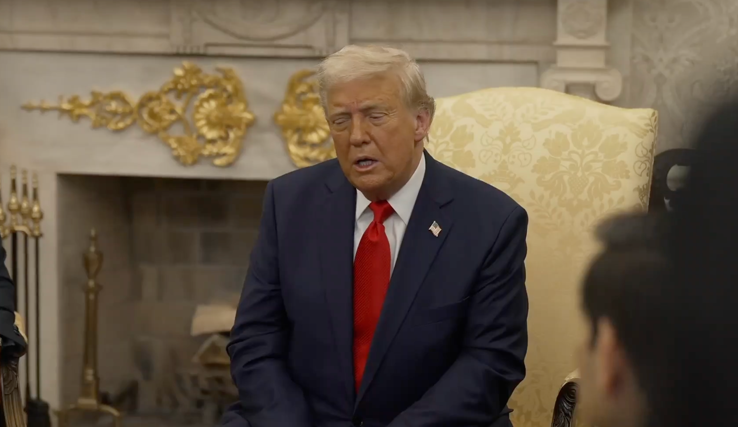 A person in a suit with a red tie sits in a formal room with ornate decor, speaking to an unseen audience
