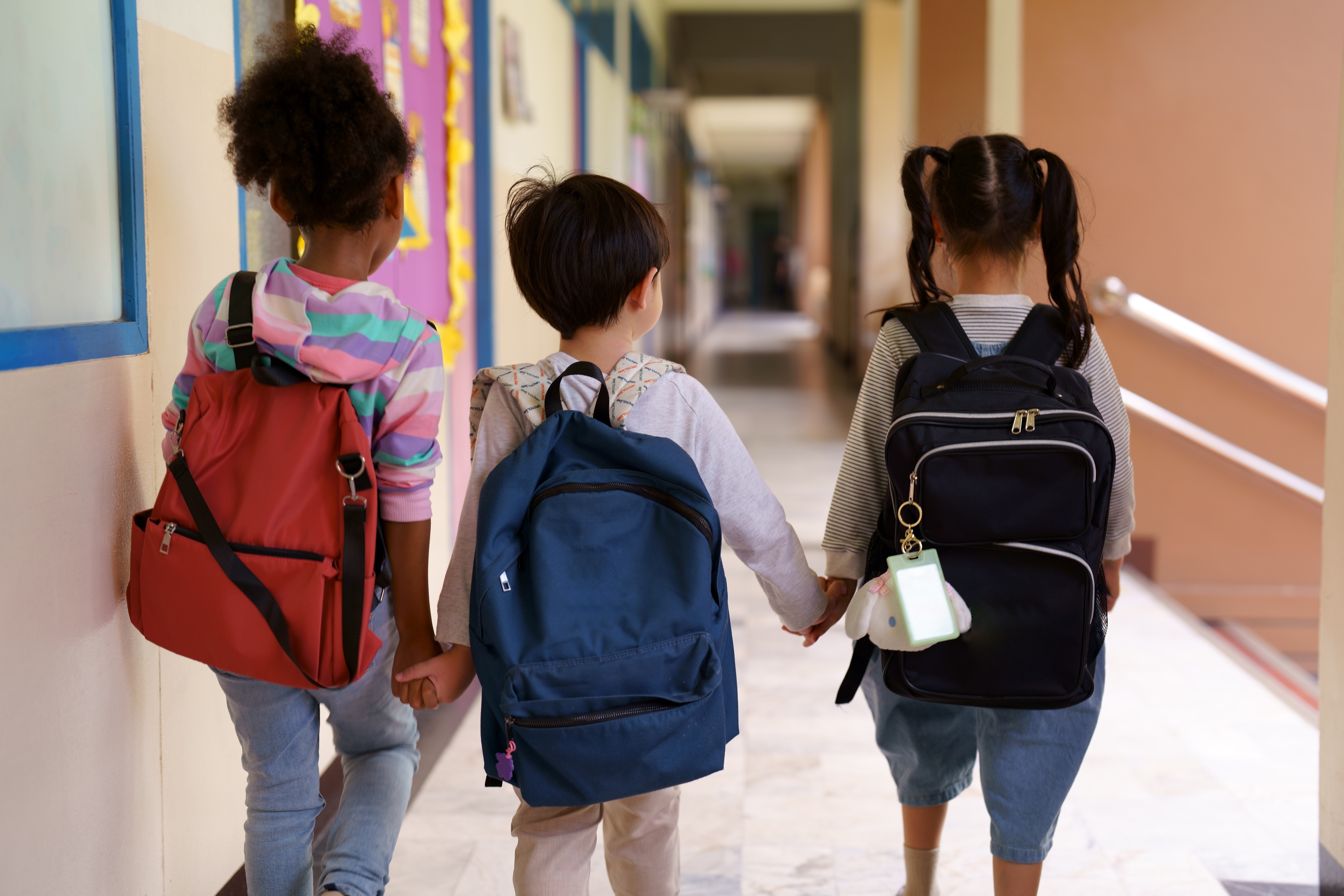 Three children with backpacks walk hand in hand down a school hallway