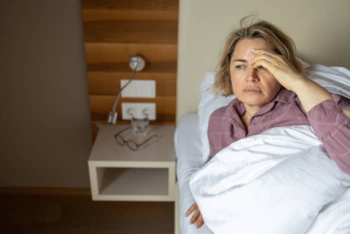 Person sitting in bed, looking contemplative, with a hand on their forehead. A bedside table with a lamp and glasses is nearby